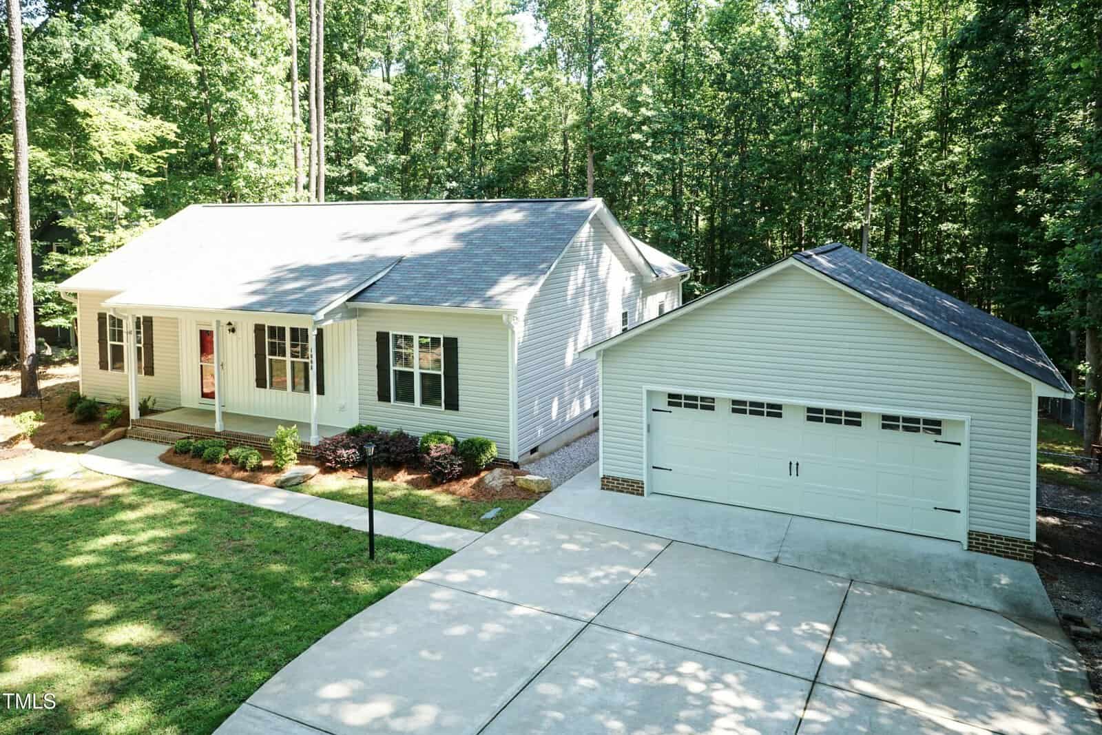 Single-story house with light gray siding, a front porch with red door, and detached two-car garage. Concrete driveway and walkway, surrounded by trees and landscaped yard with shrubs.