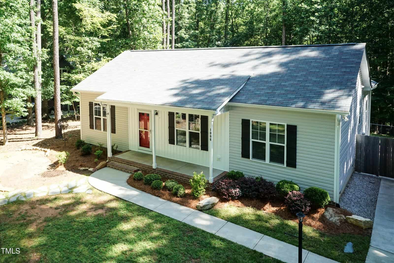 Single-story house with light-colored siding, a gray shingled roof, a red front door, black shutters, and a covered front porch. The yard has landscaped shrubs and trees, with a concrete walkway leading to the entrance.