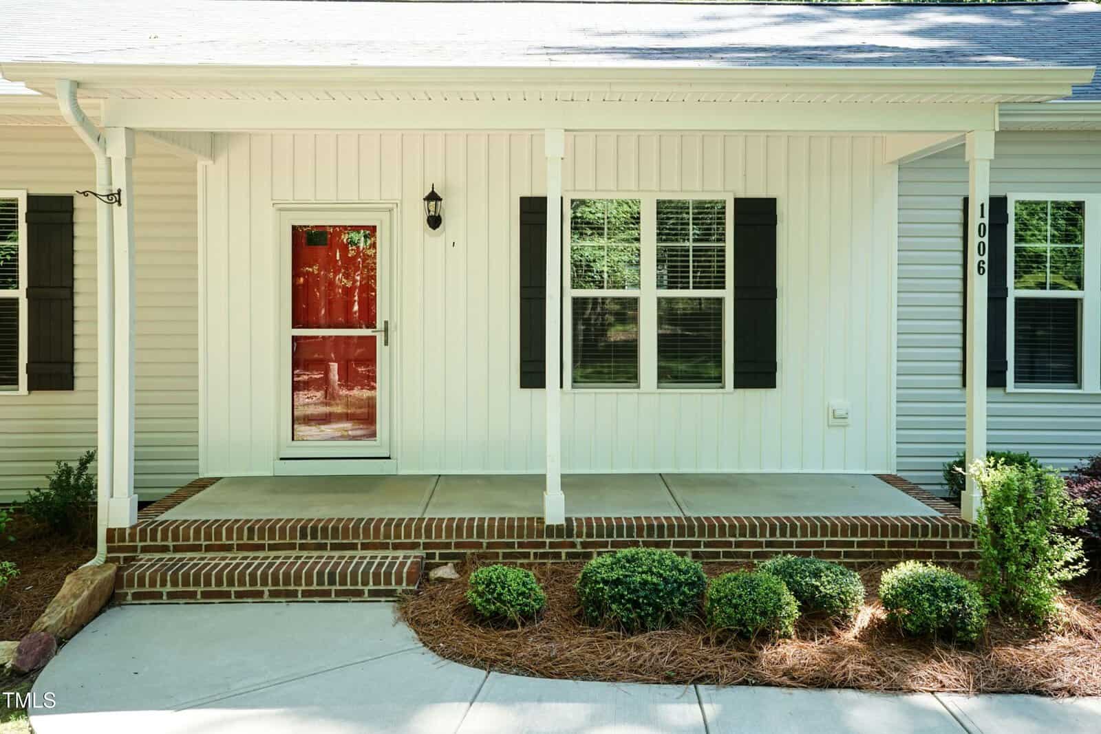A white house exterior with black shutters, a red front door with a glass storm door, a covered porch with brick steps, small shrubs, and house number 1006 displayed vertically on the wall.