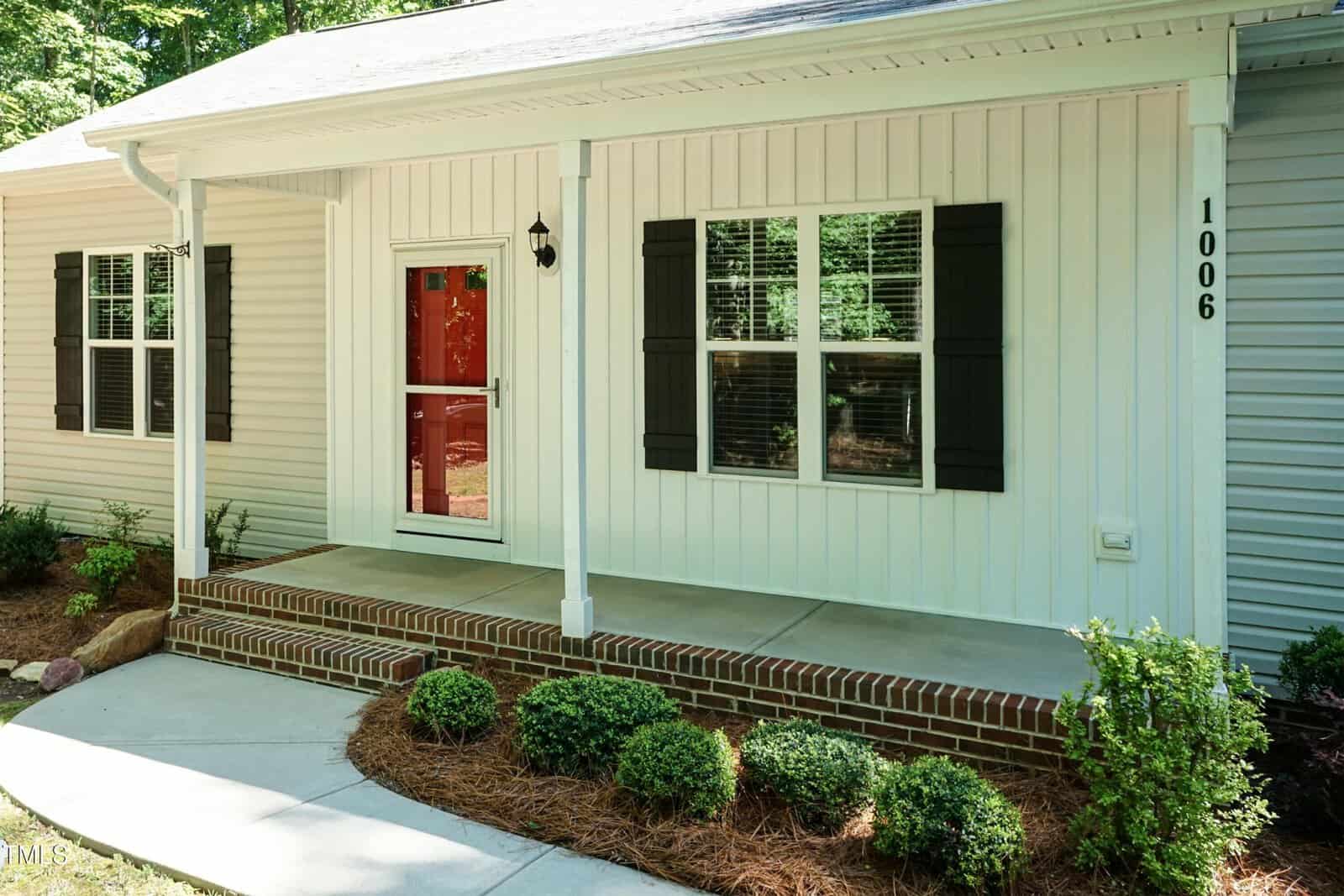 A single-story house with white siding, black shutters, a red front door, and brick steps leading to a covered front porch. The entryway is bordered by shrubs and mulch. House number 1006 is visible.