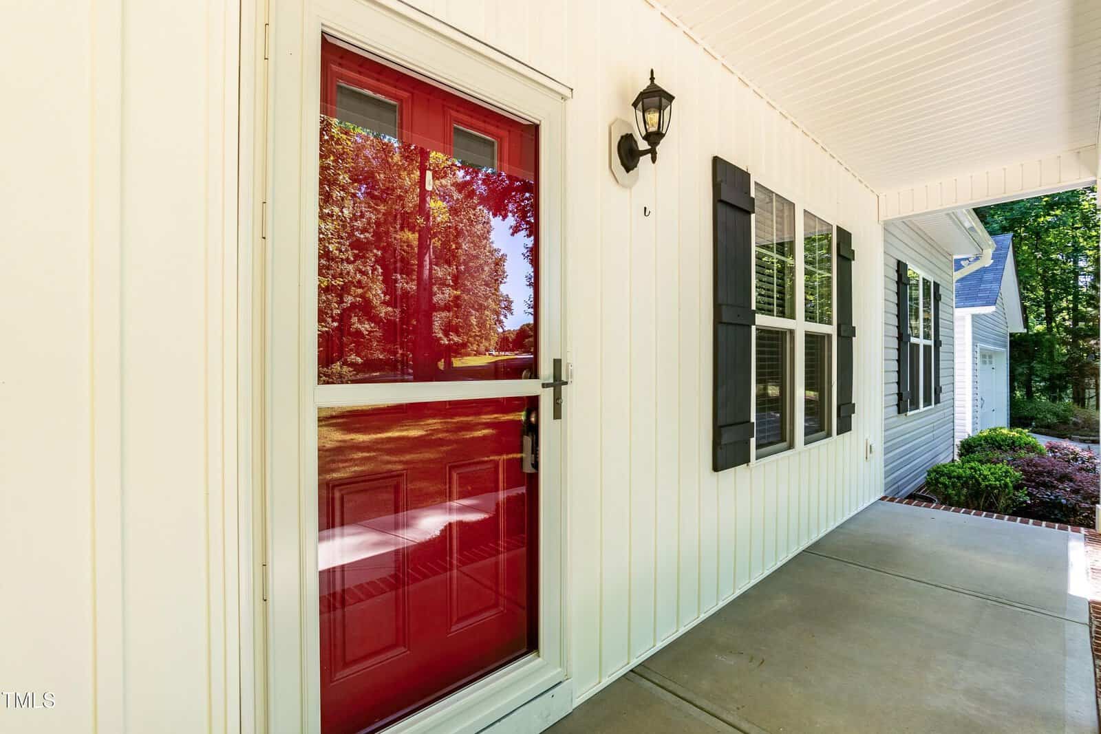 A close-up of a red front door with a glass storm door, white siding, black shutters, a wall-mounted light, and a covered concrete porch overlooking a yard with trees.