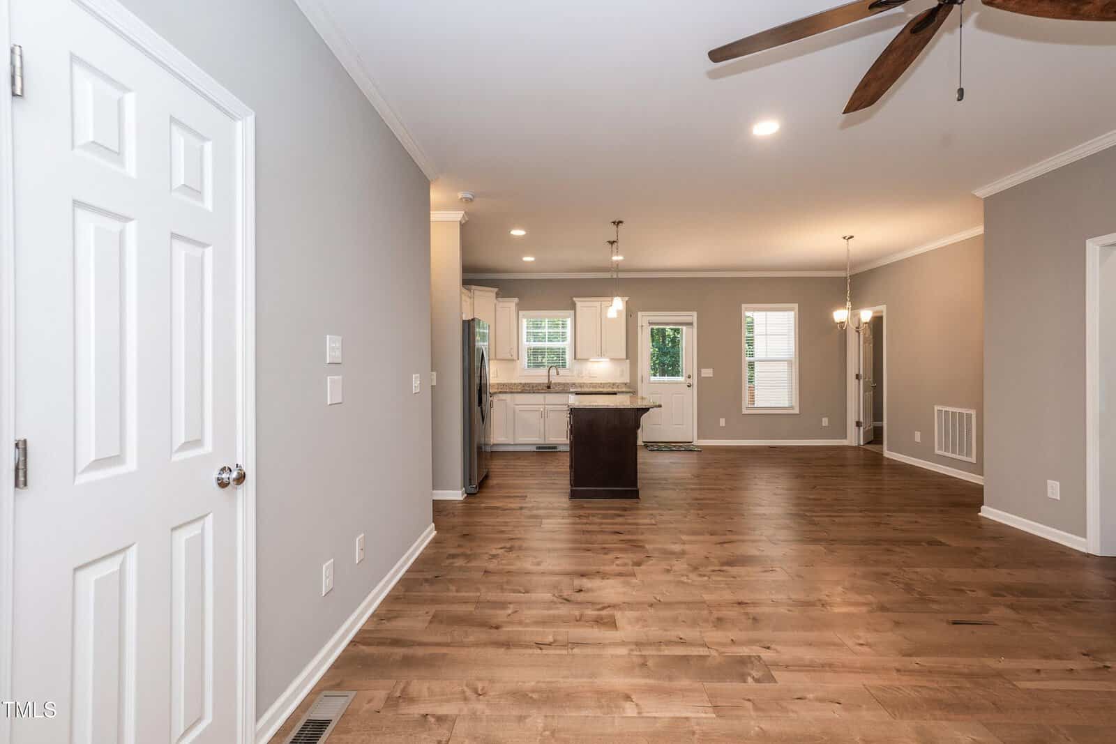 Open-concept living space with light gray walls, wood floors, ceiling fan, recessed lighting, and kitchen area in the background with white cabinets, island, and stainless steel appliances.