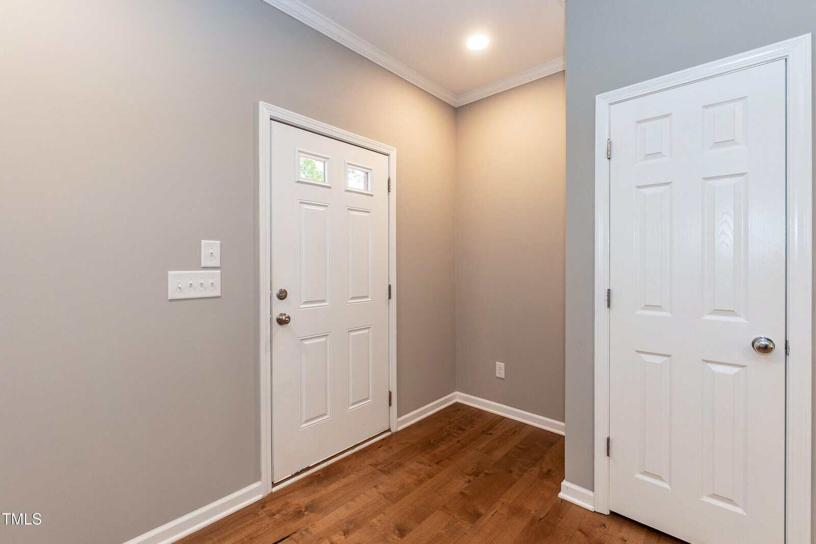 A hallway with light gray walls, wood flooring, a white front door with three square windows, and a white closet door. Ceiling light is on.