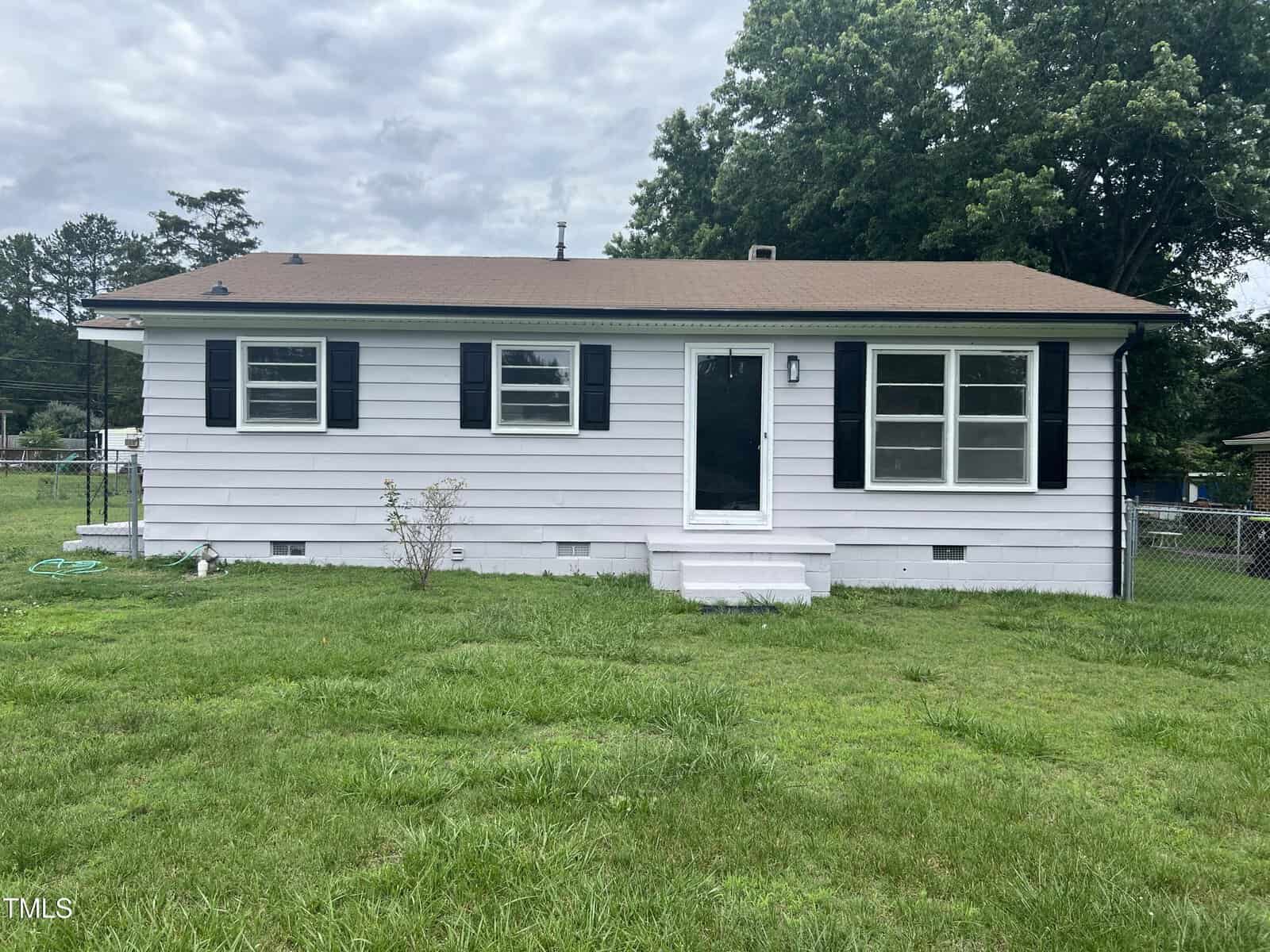 This charming, single-story house at 1851 S Clearview Drive, Henderson features white siding, black shutters, and a brown roof. A black front door with steps welcomes you; lush green grass and trees complete the inviting scene.