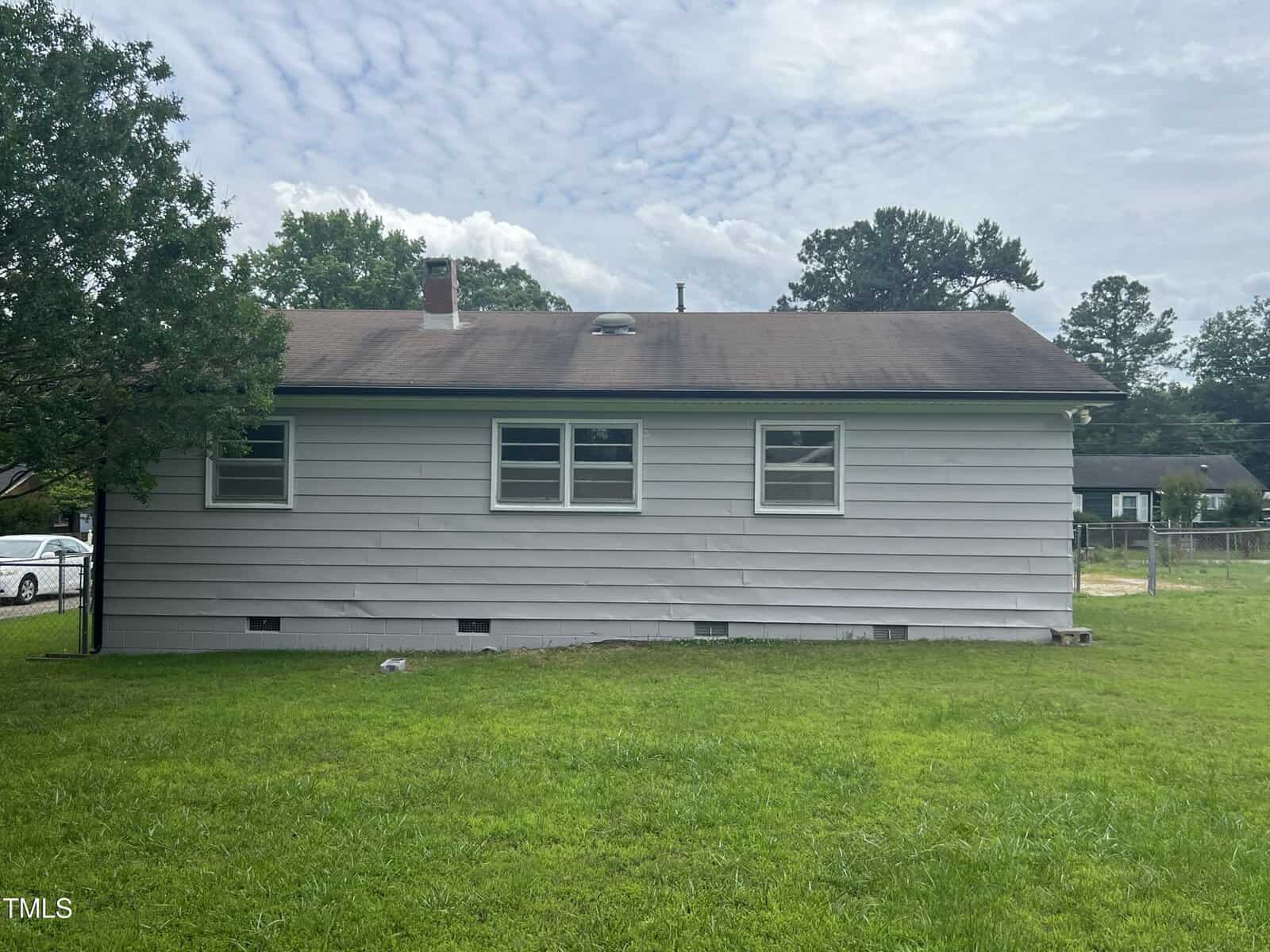 A single-story house with horizontal beige siding, three windows, and a dark shingled roof. The yard is green and grassy, with trees and other houses visible in the background.