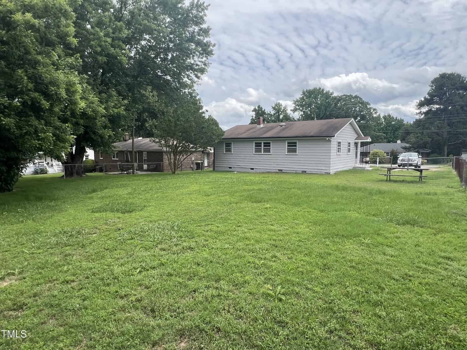 A single-story house with white siding sits on a large grassy yard. A picnic table is on the right side of the yard, and there are trees and another house nearby. The sky is partly cloudy.