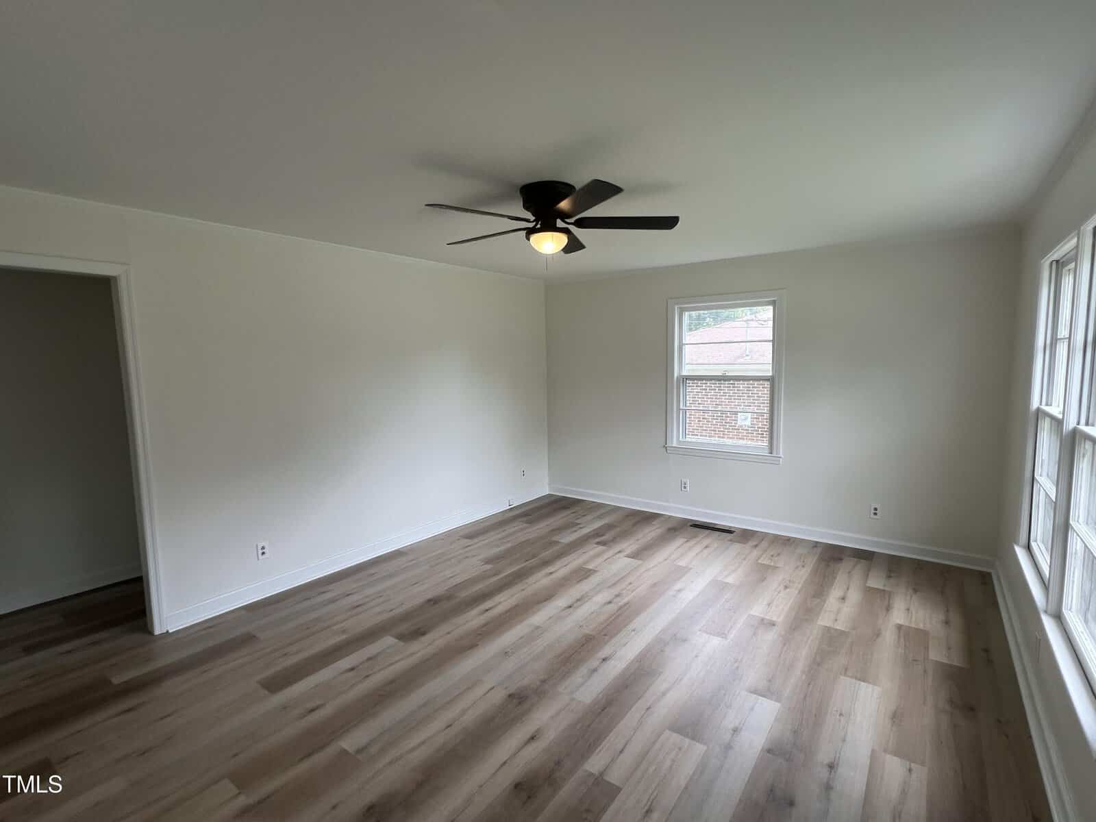 Empty room with light wood flooring, white walls, a black ceiling fan with a light, one window on the far wall, a set of three windows on the right, and a doorway on the left.