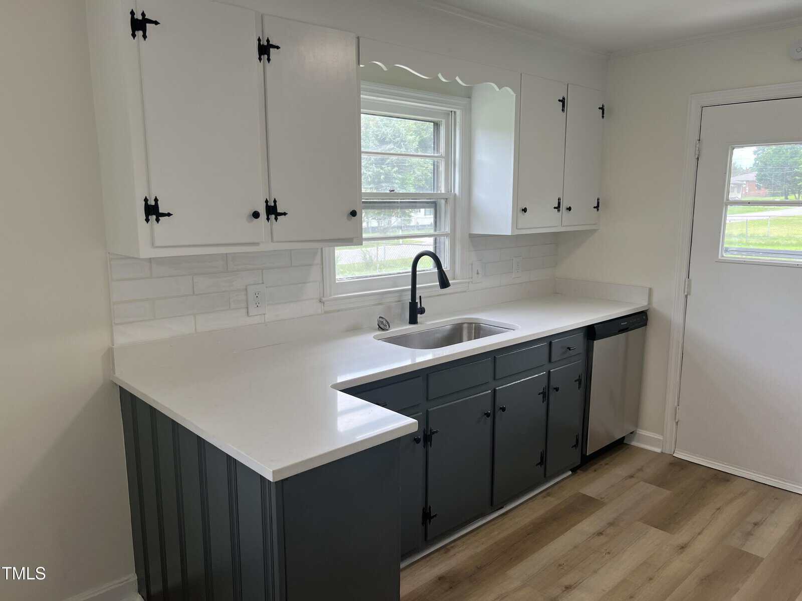 A modern kitchen with white upper cabinets, dark gray lower cabinets, a white countertop, white tile backsplash, stainless steel sink with black faucet, dishwasher, and a window above the sink.