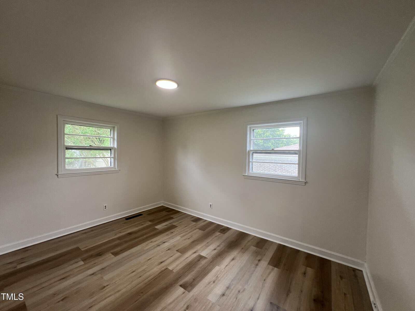 Empty room with light-colored walls, two windows with outdoor views, a ceiling light, and wood flooring. The room is well-lit by natural light from the windows.