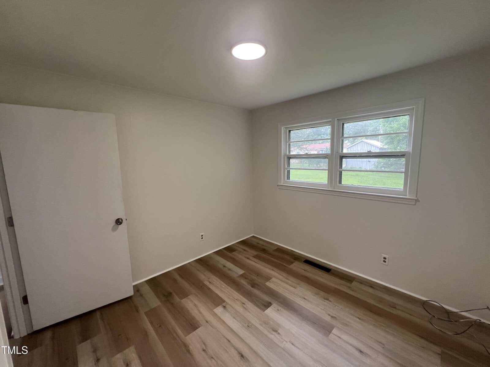 A small, empty room with light-colored walls, wood-style flooring, a closed door on the left, and two windows on the right letting in natural light.