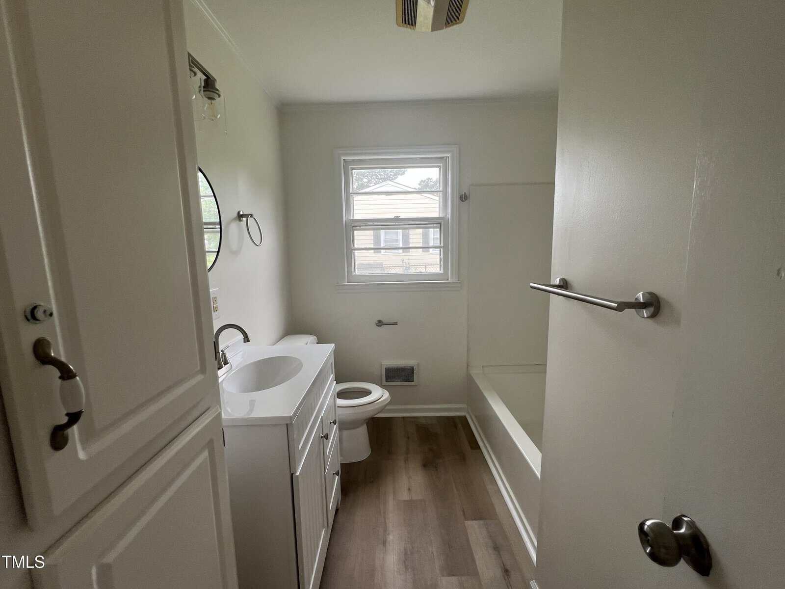 Small bathroom with white cabinets, a sink with a round mirror, toilet, bathtub with shower, grab bars, and wood-style flooring. A window lets in natural light above the toilet.