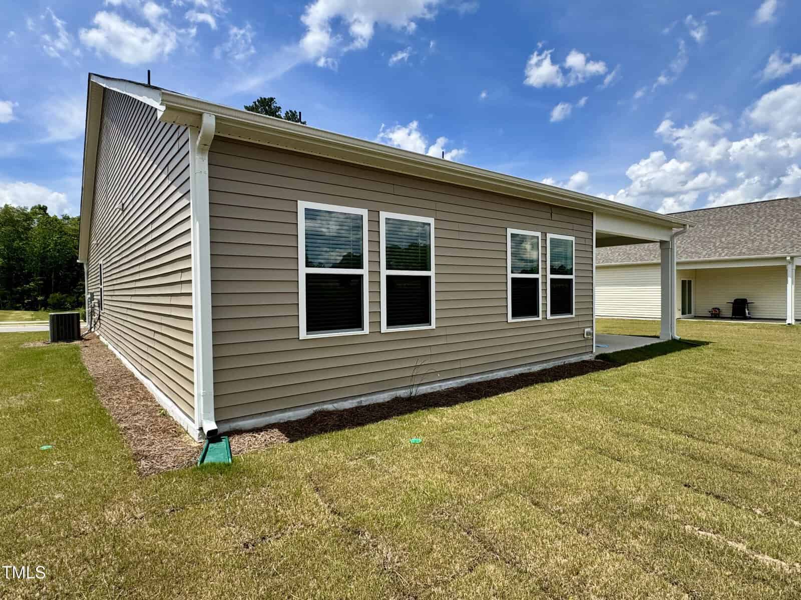 Single-story house with tan siding, three double windows, and white trim; surrounded by a green lawn under a blue sky with scattered clouds. A second house is visible in the background.