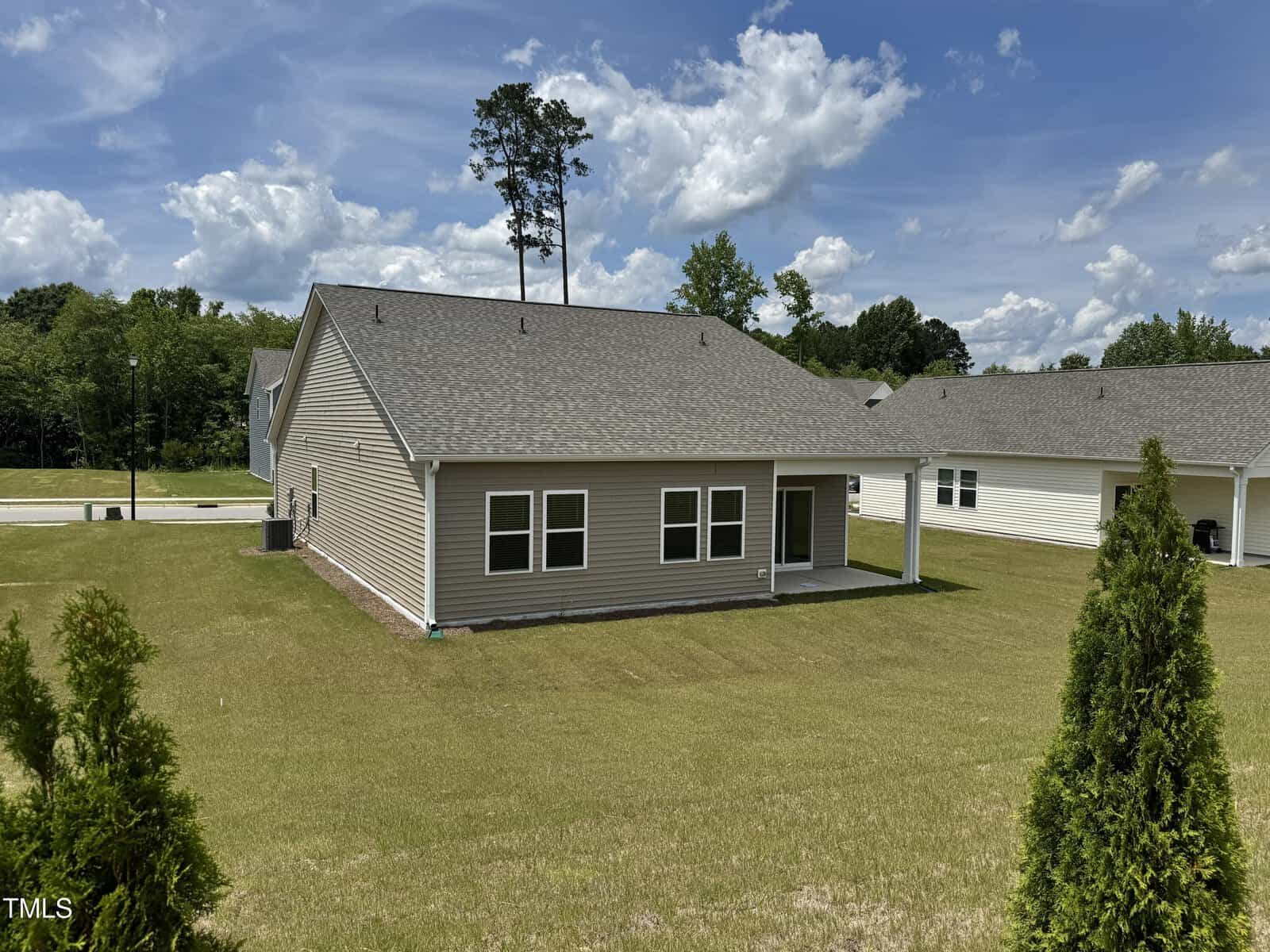 A single-story beige house with a gray roof and covered back patio sits on a large, green lawn under a partly cloudy sky. Another similar house is visible to the right, with trees in the background.