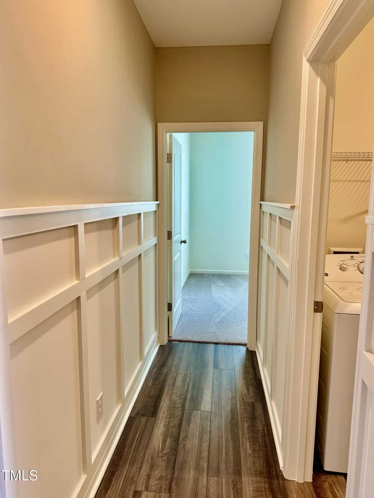 A hallway with beige walls and white wainscoting, leading to a carpeted room. To the right, there is a partially visible laundry area with a washer and wire shelf. The floor is dark wood-patterned vinyl or laminate.