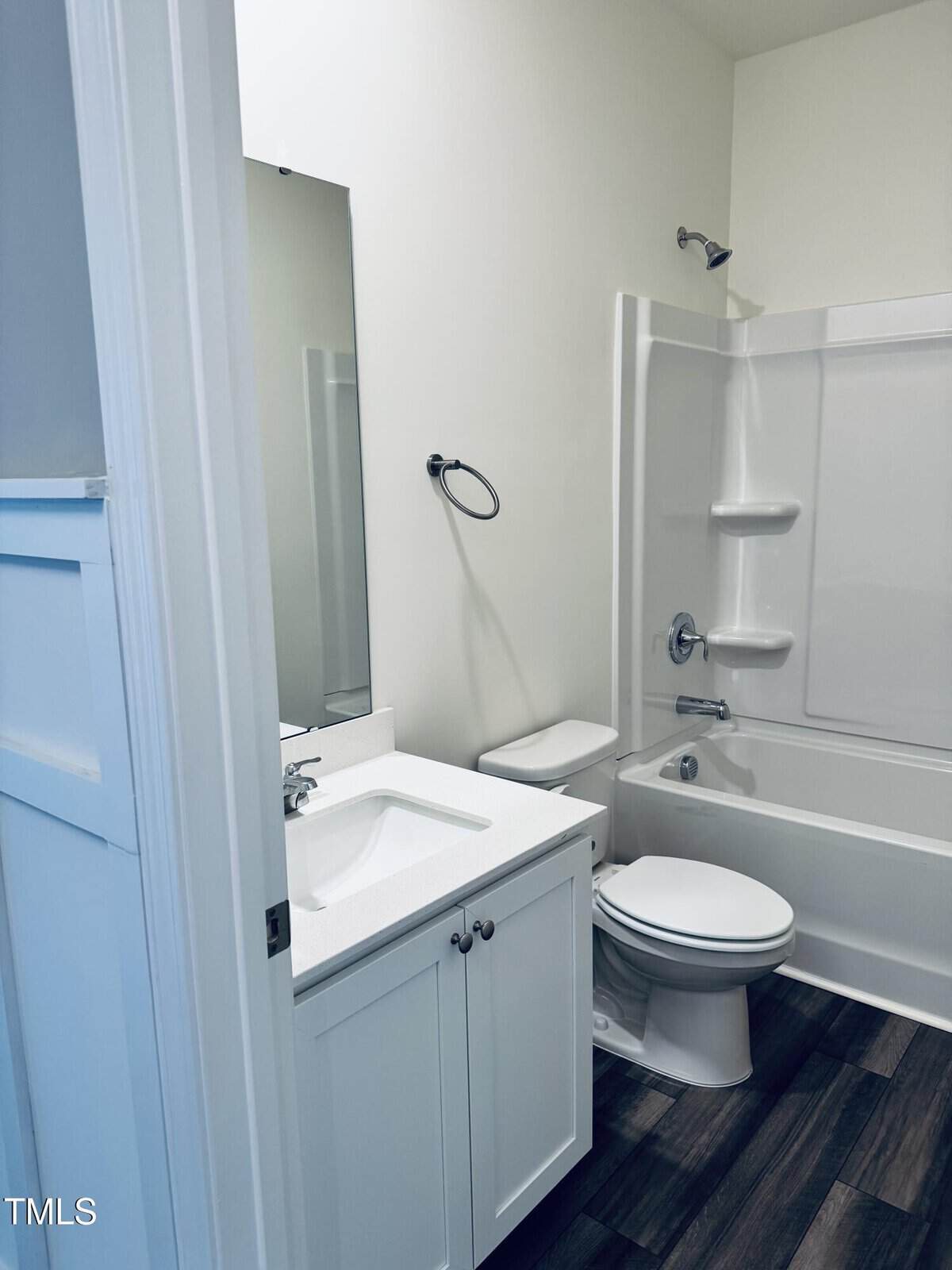 A modern bathroom with white walls, a white vanity and sink, a toilet, a bathtub-shower combo with shelves, a large mirror, and dark wood-look flooring.