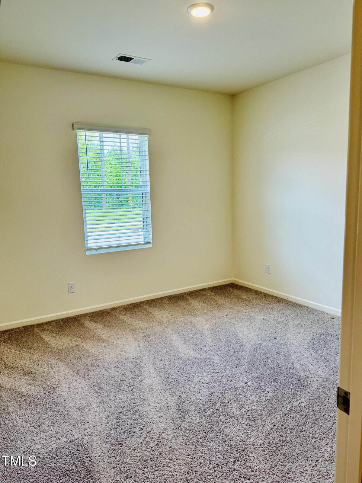 Empty room with beige carpet, off-white walls, a single window with blinds letting in natural light, and a ceiling light fixture. The room appears clean and well-lit.