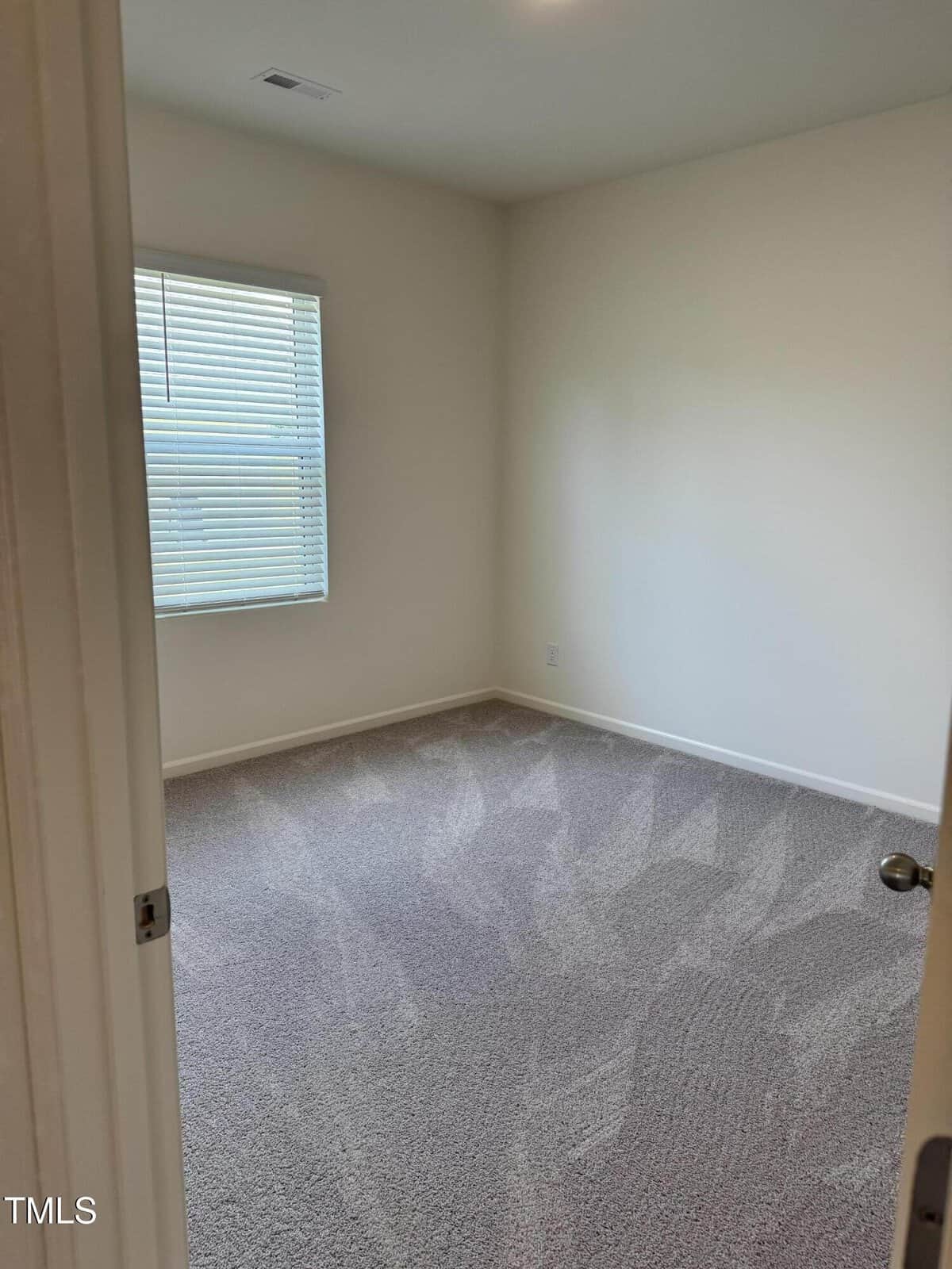 Empty bedroom with beige carpet, cream-colored walls, a single window with blinds, and a ceiling vent. The room is clean and unfurnished, with sunlight filtering through the window.