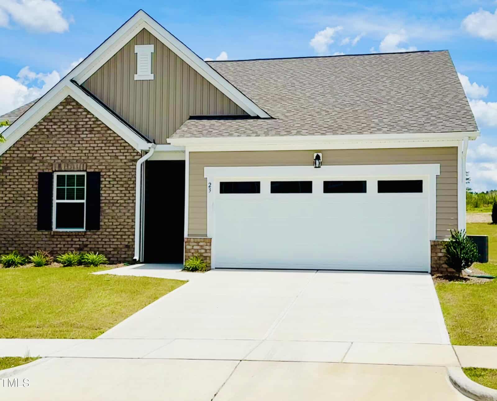 A single-story suburban house with a brick and beige siding exterior, a gabled roof, black shutters, attached two-car garage with white doors, and a concrete driveway under a blue sky with clouds.