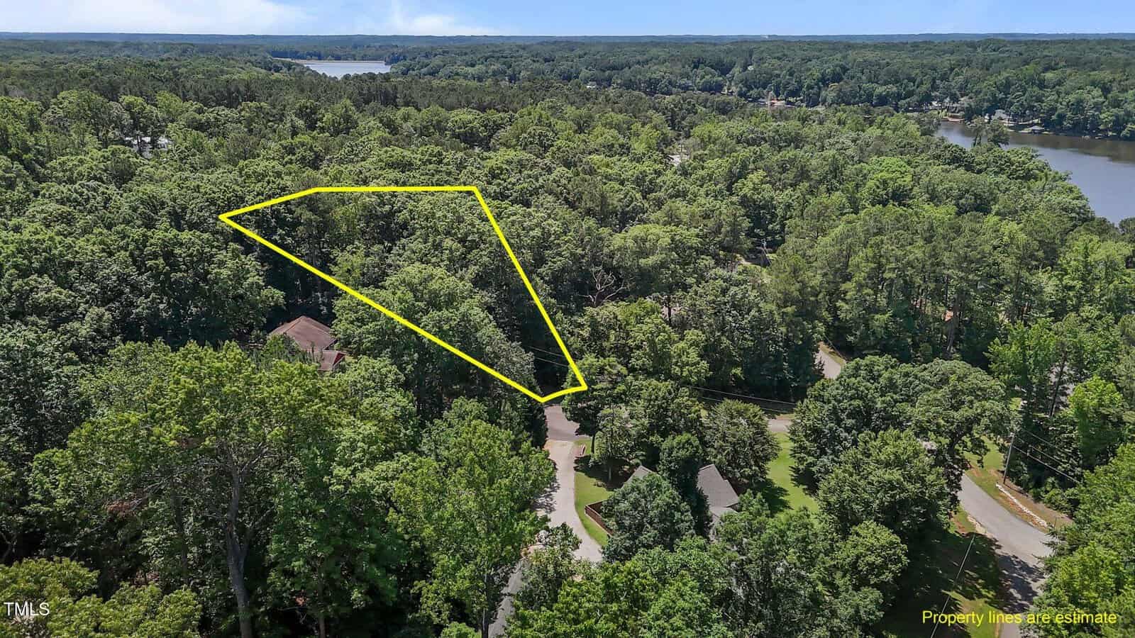 Aerial view of a wooded area with a triangular plot of land outlined in yellow, surrounded by dense trees, nearby houses, and roads; a lake is visible in the background.