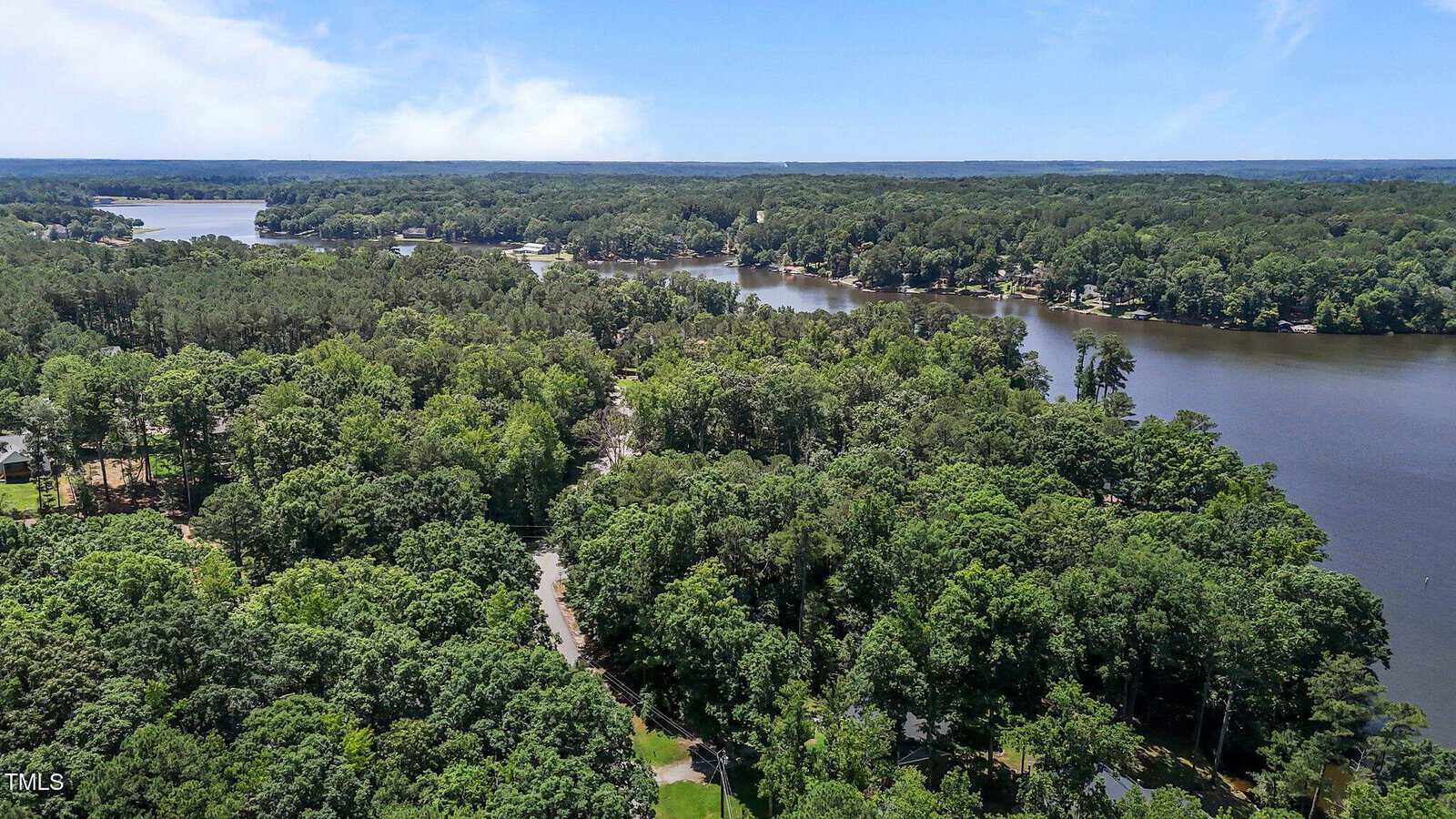 Aerial view of a wooded area with dense green trees surrounding a winding lake. Several houses are partially visible among the trees, and the sky is mostly clear with some clouds in the distance.