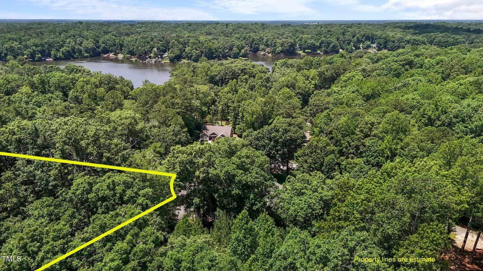 Aerial view of a house surrounded by dense trees near a lake; a yellow outline marks the property boundary in the wooded area. Blue sky is visible above the forested landscape.