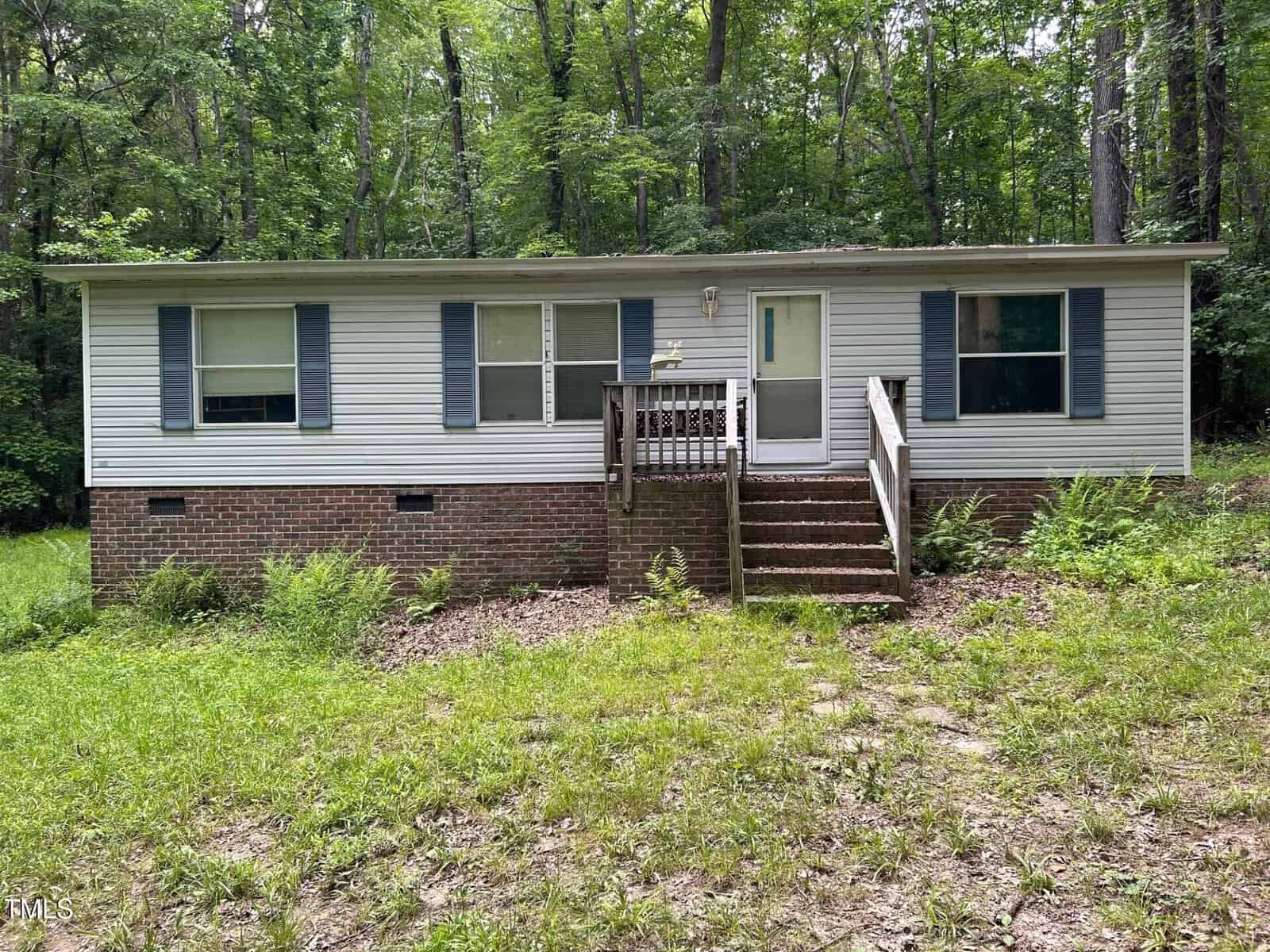 A single-story house with white siding, brick foundation, blue shutters, and a small front porch with steps, nestled in a wooded area with overgrown grass at 3539 Charlies Court in Wake Forest.