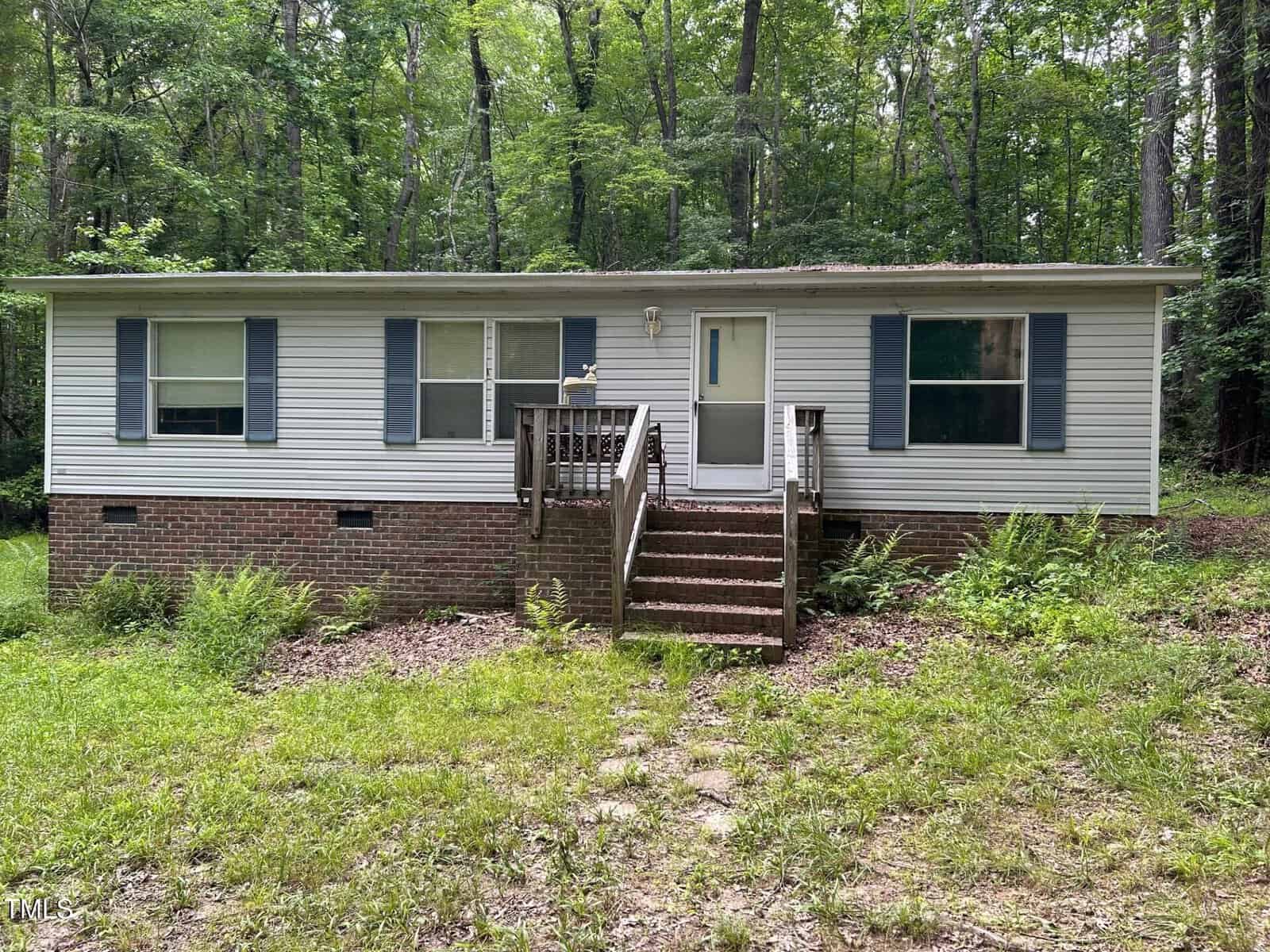A single-story house with white siding, blue shutters, and a brick foundation stands in front of a wooded background. A small porch with wooden steps leads to the front door, and the yard is overgrown with grass and ferns.