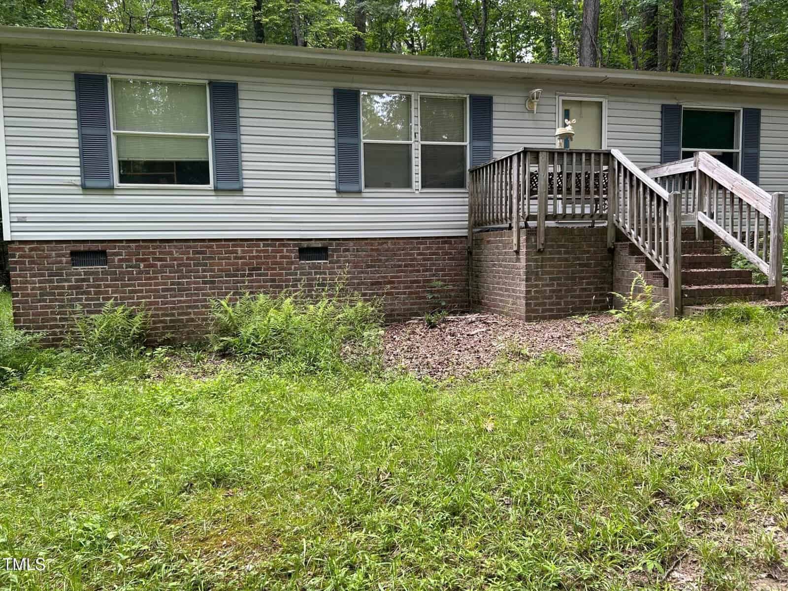 A single-story house with white siding, blue shutters, and a brick foundation. A wooden staircase with railings leads to the front door. The yard has overgrown grass and some weeds near the house.