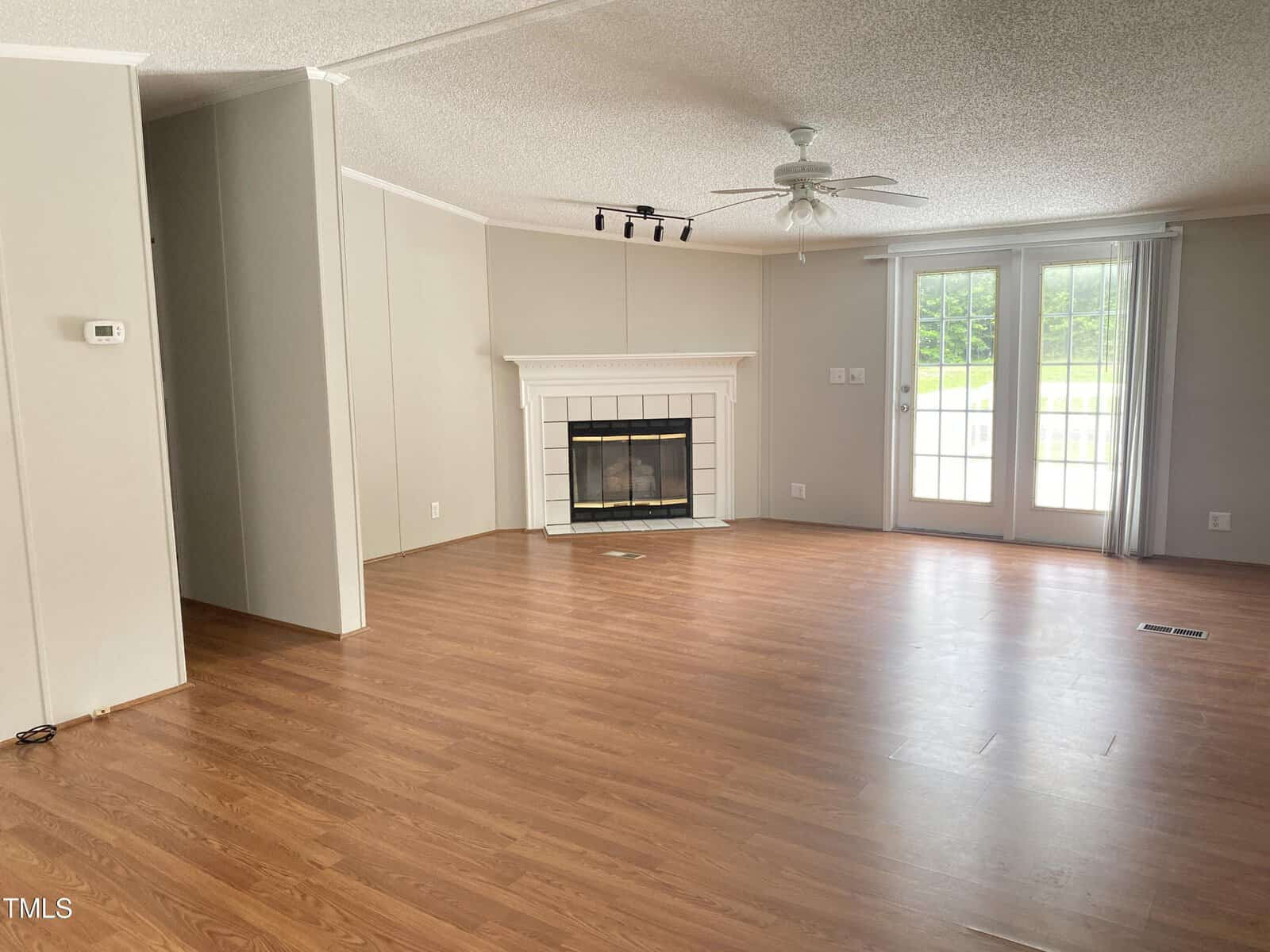 Unfurnished living room with wood flooring, a white fireplace, ceiling fan, beige walls, and double glass doors leading outside. There is a small track light above the fireplace and natural light entering the room.