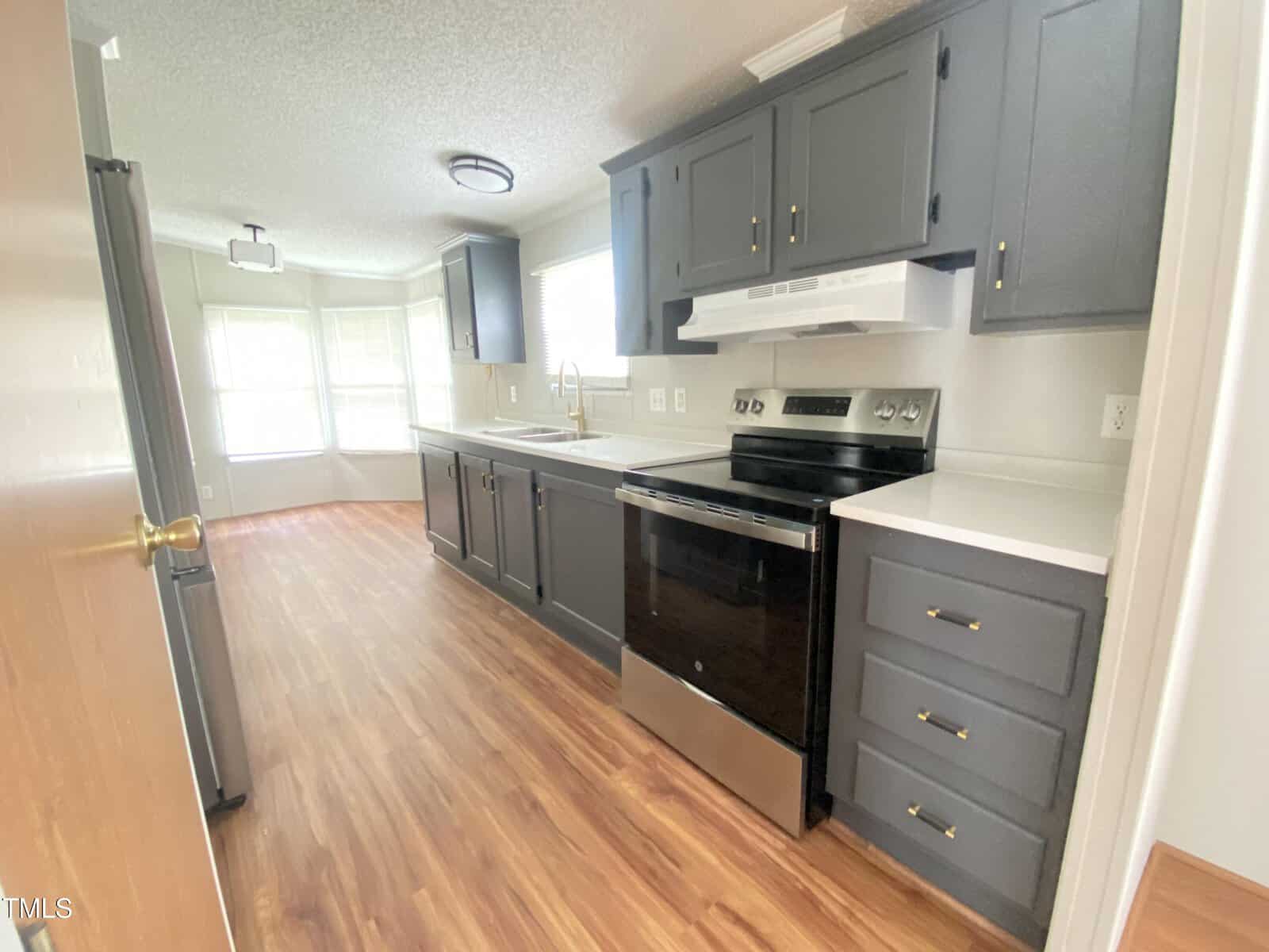 A modern kitchen with dark gray cabinets, white countertops, stainless steel oven, and wood-style flooring. Large windows in the adjacent dining area provide natural light.