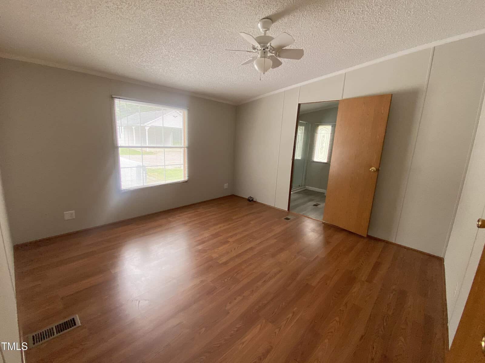 An empty room with wood flooring, light-colored walls, a ceiling fan, a large window with blinds, and two wooden doors, one partially open showing an adjacent room. Natural light comes in through the window.