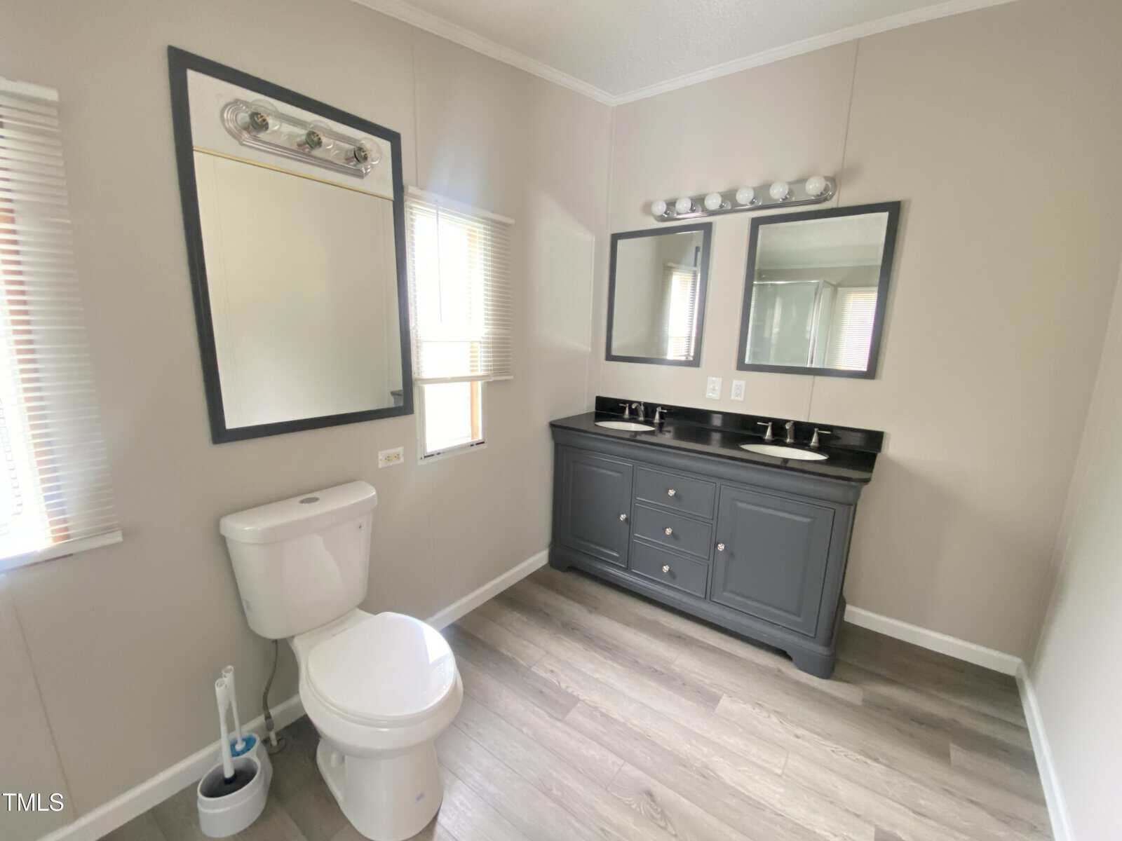 A bathroom with light-colored walls, a white toilet, a cleaning brush, a large mirror above the toilet, two windows, and a double-sink vanity with two mirrors and overhead light fixtures. The floor is wood-patterned.