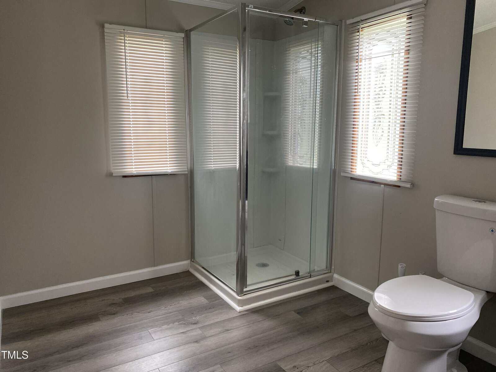 A bathroom with a corner glass shower, a white toilet, two windows with blinds, light-colored walls, and wood-look flooring.