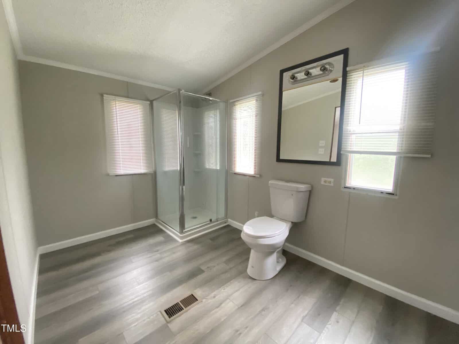 A bathroom with light gray walls and flooring, featuring a glass shower enclosure, a toilet, a large wall mirror, and three windows with blinds that let in natural light.