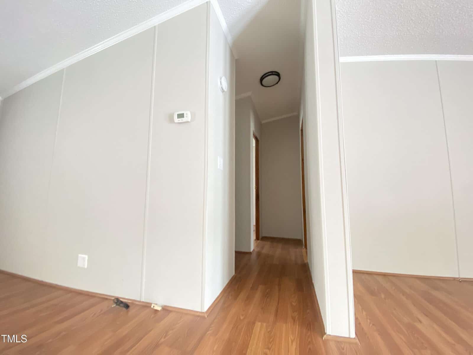 A hallway with light-colored walls and wood laminate flooring, featuring a visible thermostat, an overhead light, and a partially visible corner of an adjacent room.