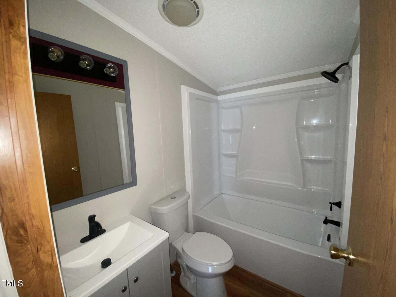 Small bathroom with a white bathtub-shower combo, white toilet, and white sink vanity. There is a mirror above the sink, wood flooring, and a wooden door partly open.