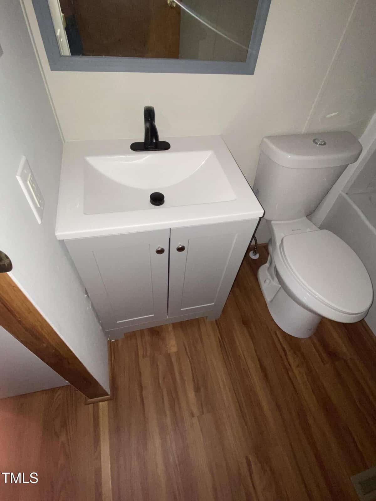 Small bathroom with a white vanity sink, black faucet, and a toilet beside it. The floor is wood-patterned, and there’s part of a mirror above the sink.