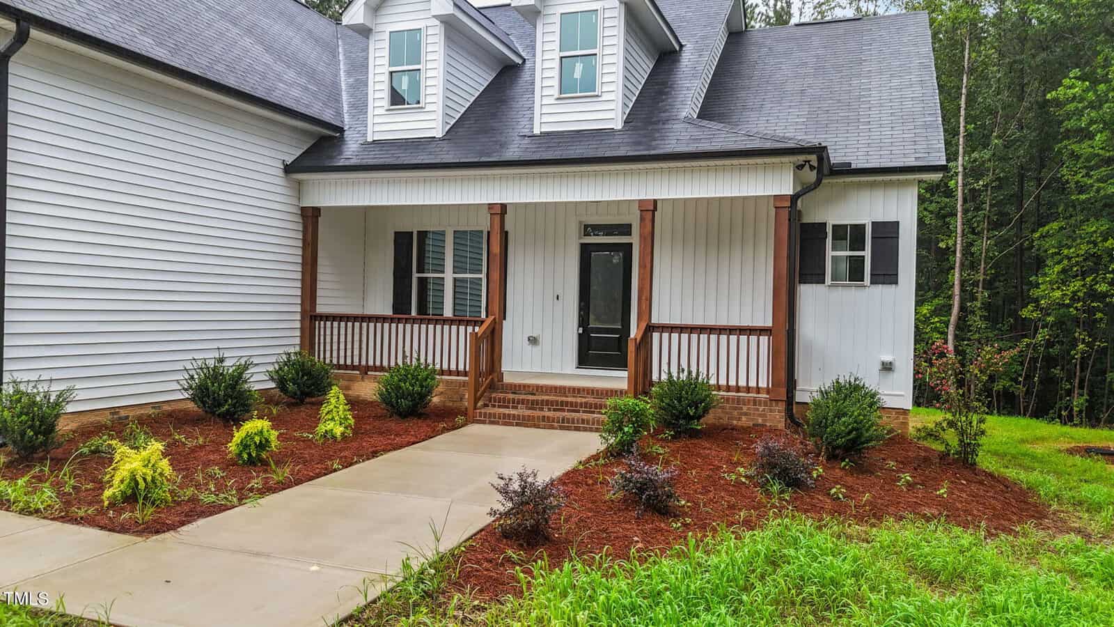 A white house with a covered front porch, brown wooden railings, and steps leading to the entrance. Bushes and small plants are arranged along a paved walkway in a mulched front yard. Trees are visible in the background.