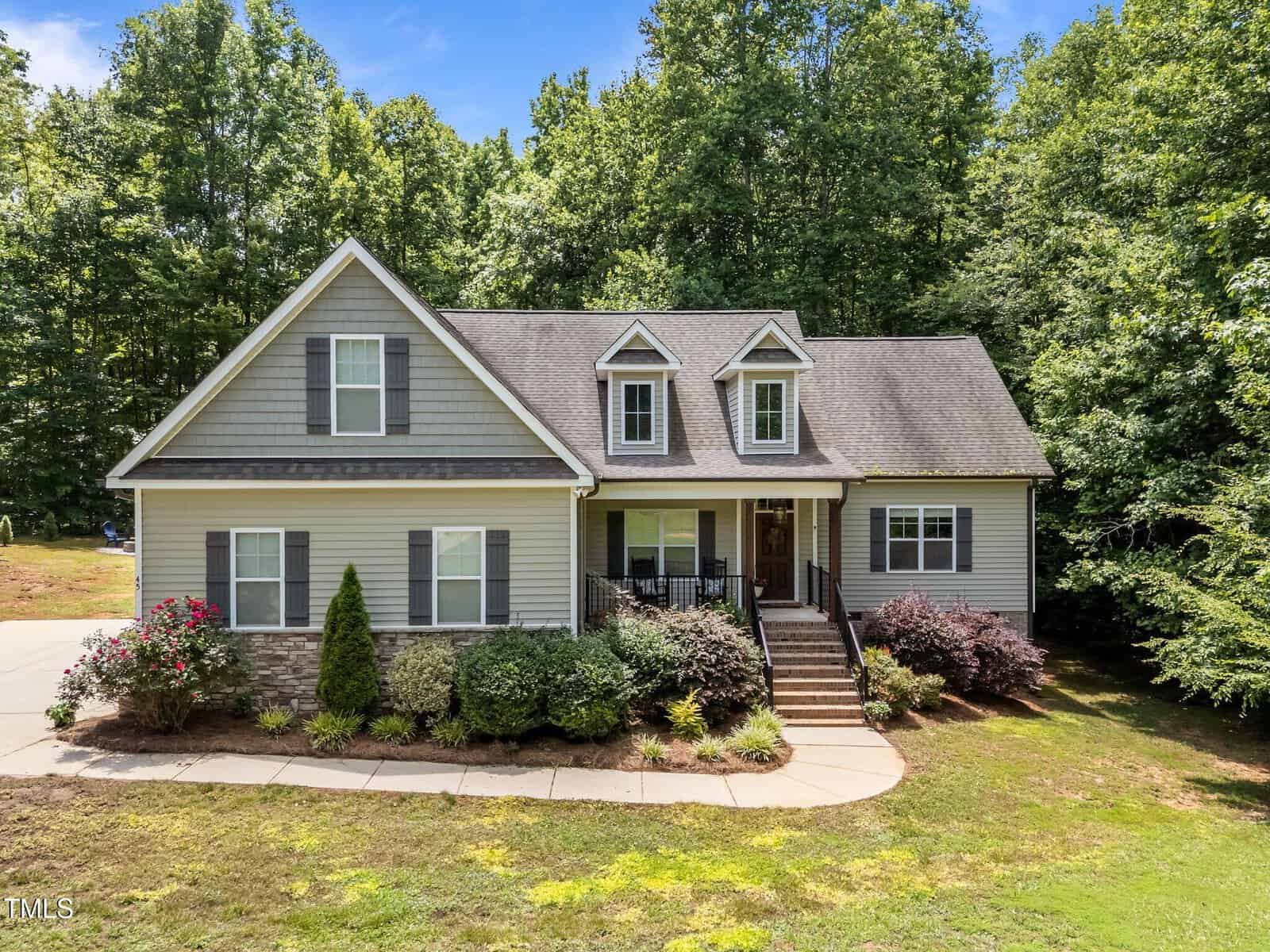 Charming two-story gray house with white trim, gabled roof, and front porch with steps located at 45 Chaucer Court W, Zebulon. Surrounded by bushes, small trees, and a curved sidewalk, this lovely residence features a driveway visible to the left and a peaceful forested area in the background.