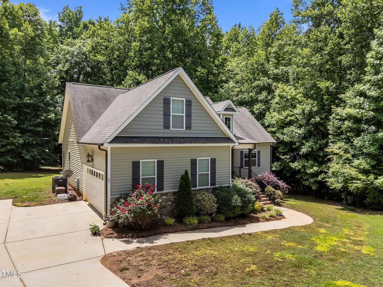 A gray two-story house with white trim, a gable roof, an attached garage, and a small front porch. The yard features shrubs and flowers, and is surrounded by tall, green trees under a blue sky.