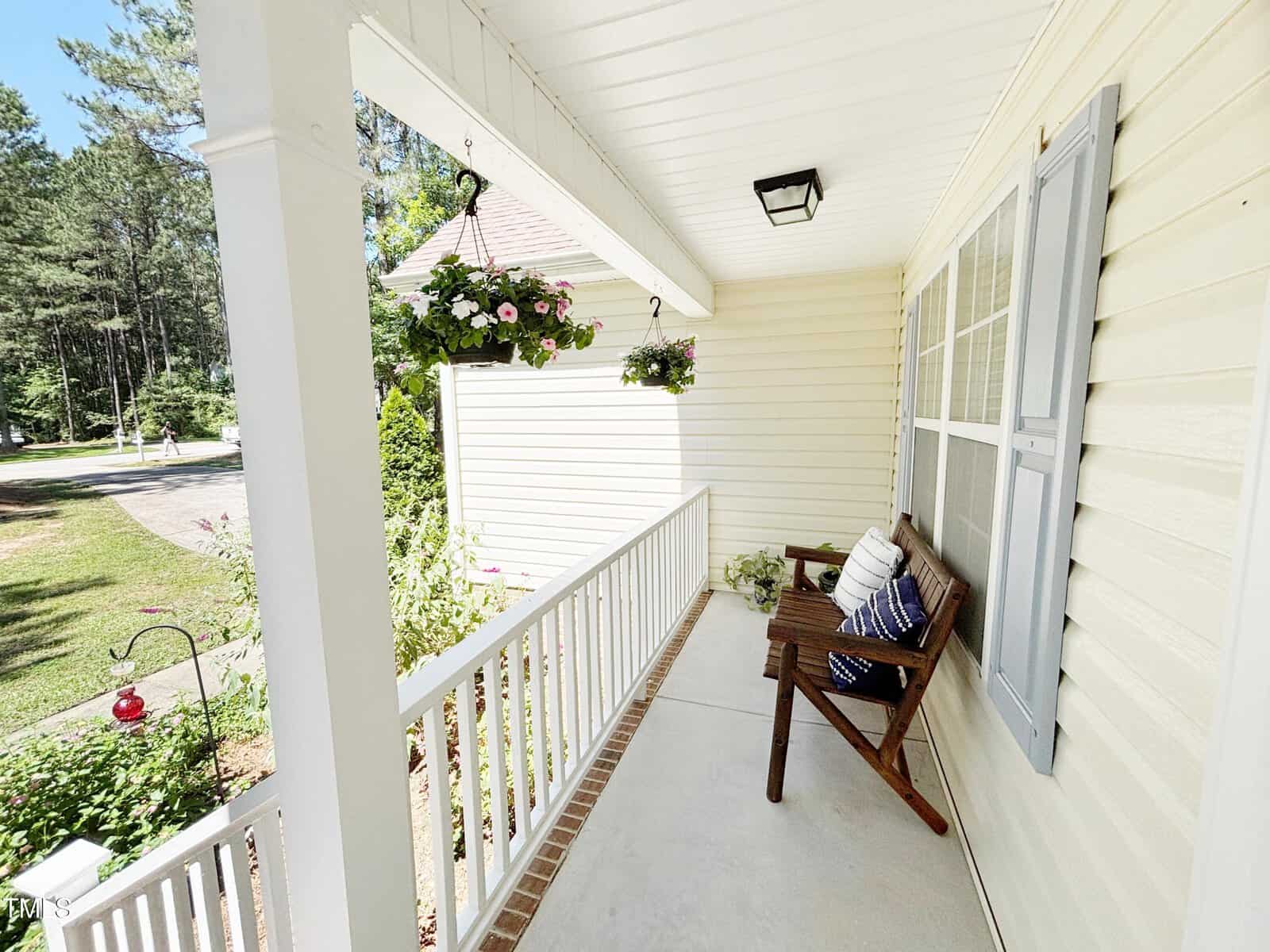 A cozy front porch with a wooden bench, blue and white pillows, potted flowers, hanging baskets, white railing, and cream siding, overlooking a green yard and trees on a sunny day.
