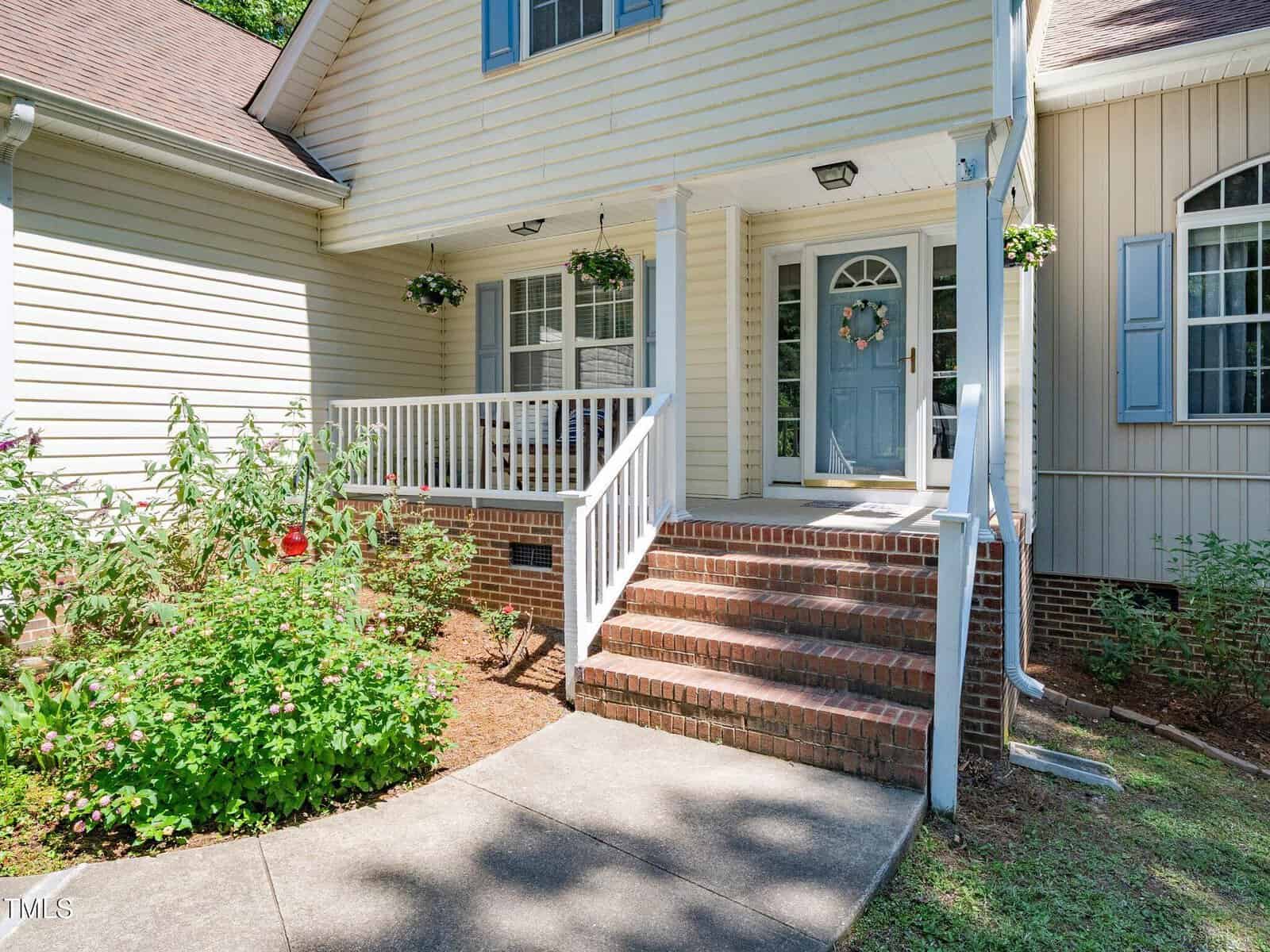 Front entry of a house with light siding, a brick staircase, and a porch with hanging plants. Blue front door has a wreath and windows on each side. Flowering plants grow near the walkway and stairs.