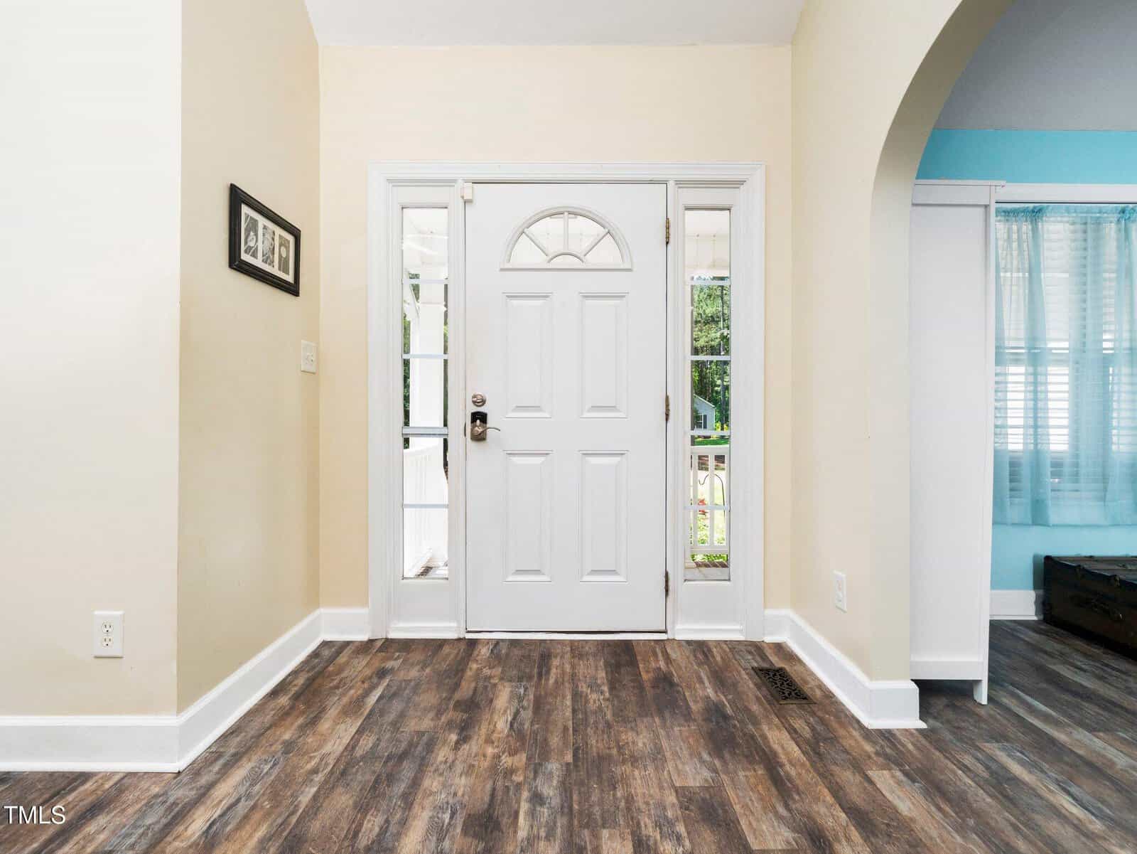 A white front door with an arched window is centered in a room with wood-look flooring. To the left, a small framed picture hangs on the wall, and to the right, an arched doorway leads to a room with blue walls and white curtains.