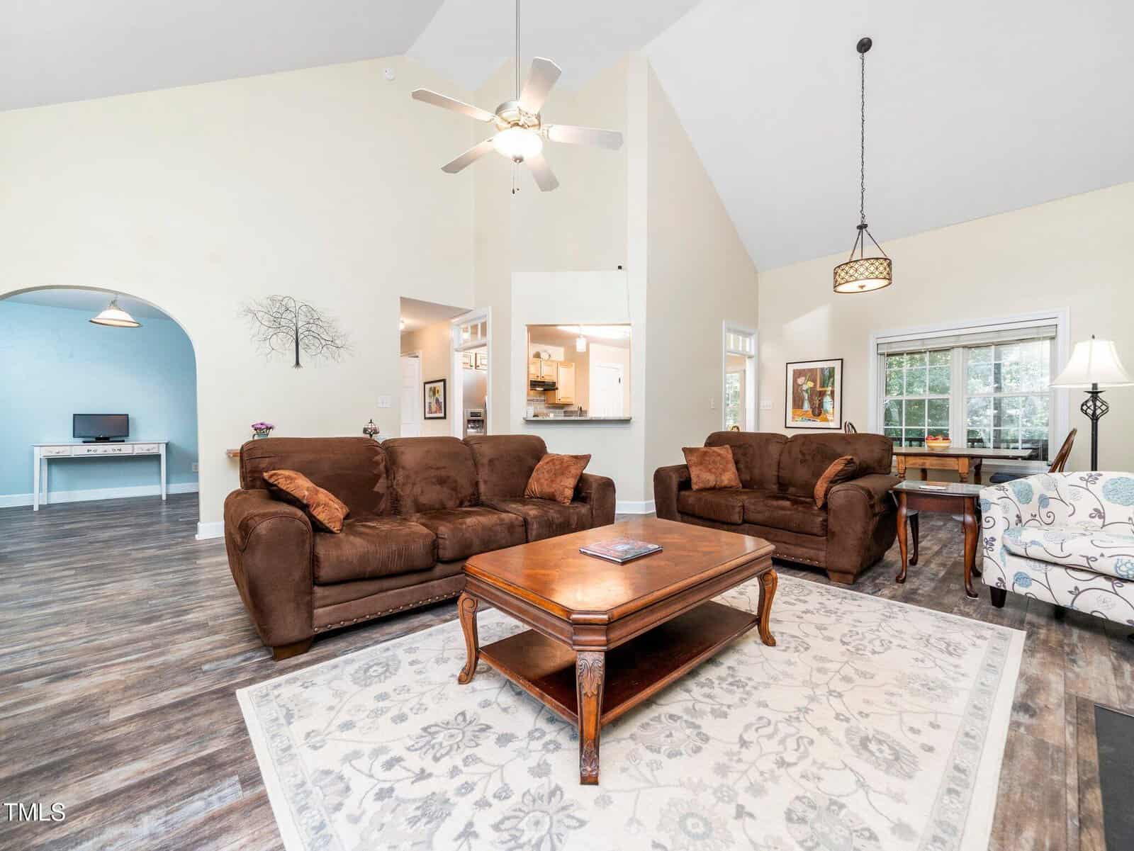 Living room with brown sofas, a wooden coffee table on a patterned rug, a ceiling fan, and large windows. An archway leads to a desk area; an open wall shows the kitchen. The floor is wood with a gray tone.