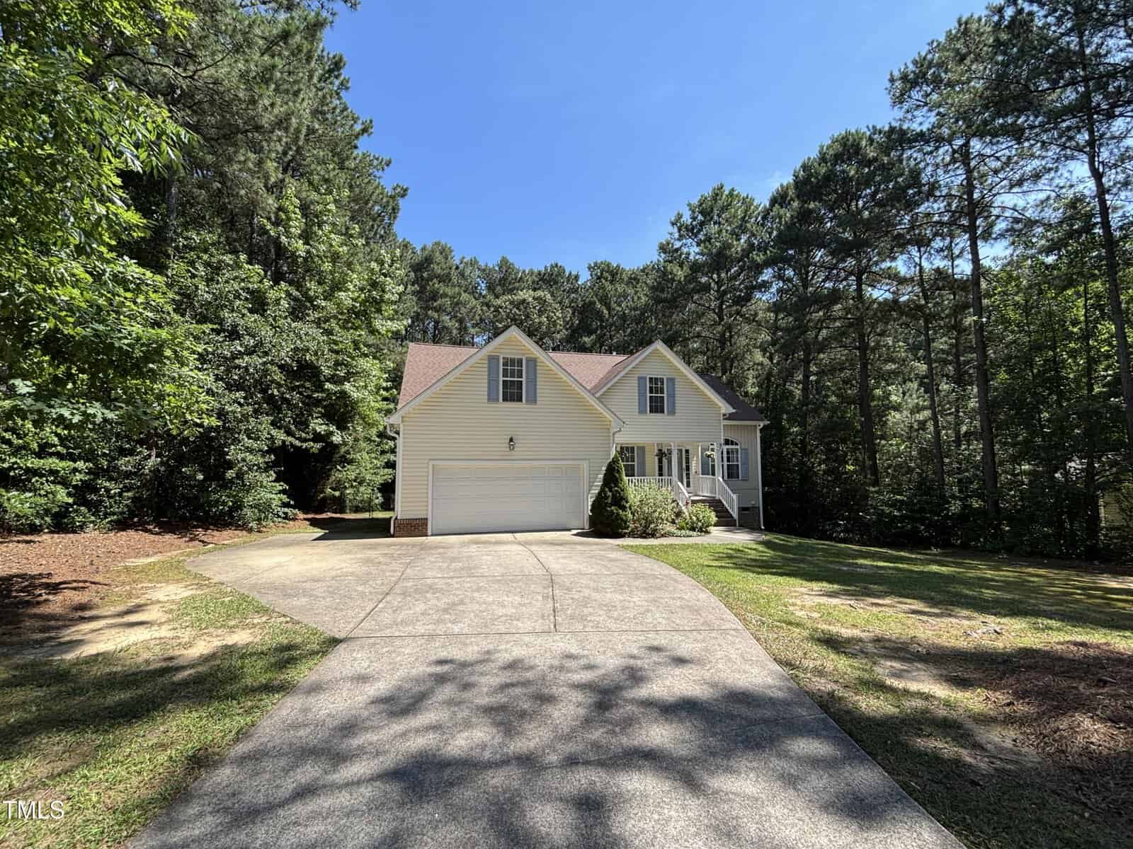 A two-story light yellow house with a double garage and gabled roof sits at the end of a long driveway, surrounded by tall pine trees and shaded lawn, on a sunny day.