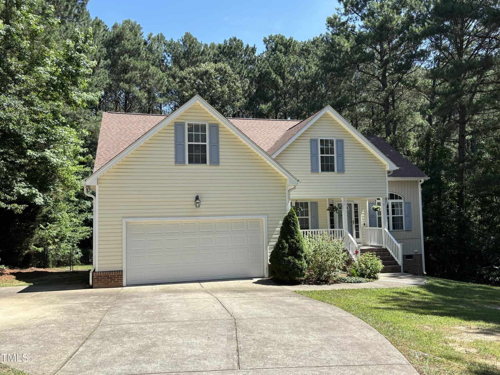 A light yellow, two-story suburban house with a gable roof, attached double garage, front porch with white railings, and a concrete driveway, surrounded by green trees and bushes under a clear blue sky.