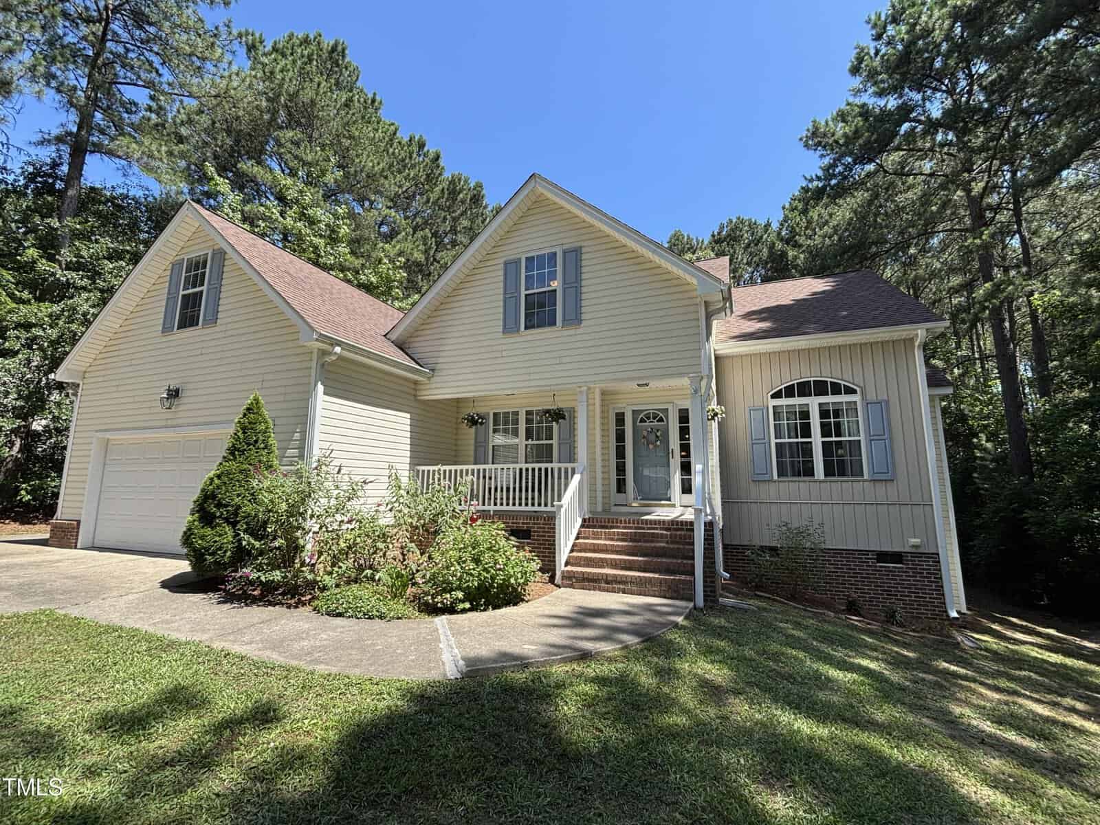 A light beige two-story house with a front porch, brick steps, and a gabled roof sits surrounded by trees and greenery under a clear blue sky. A curved driveway leads to an attached garage on the left.