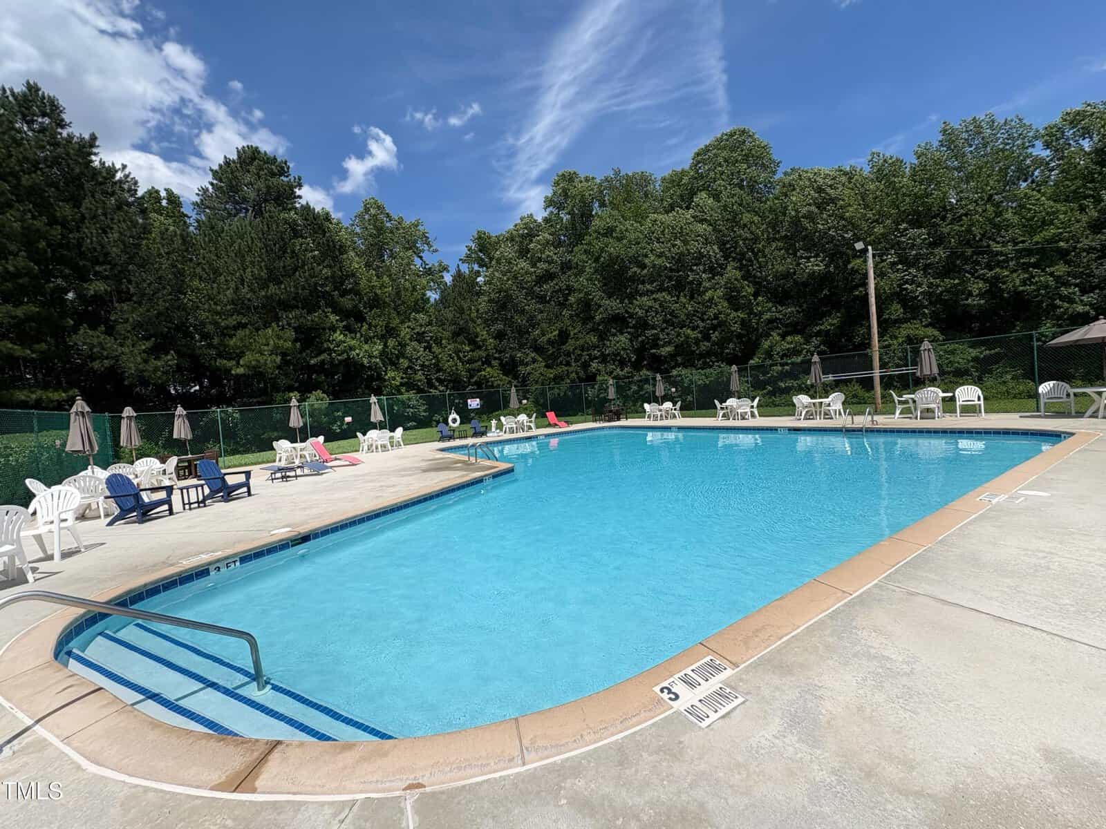 Outdoor swimming pool with clear blue water surrounded by lounge chairs, tables, and umbrellas. Green trees and a fence line the background under a partly cloudy sky.