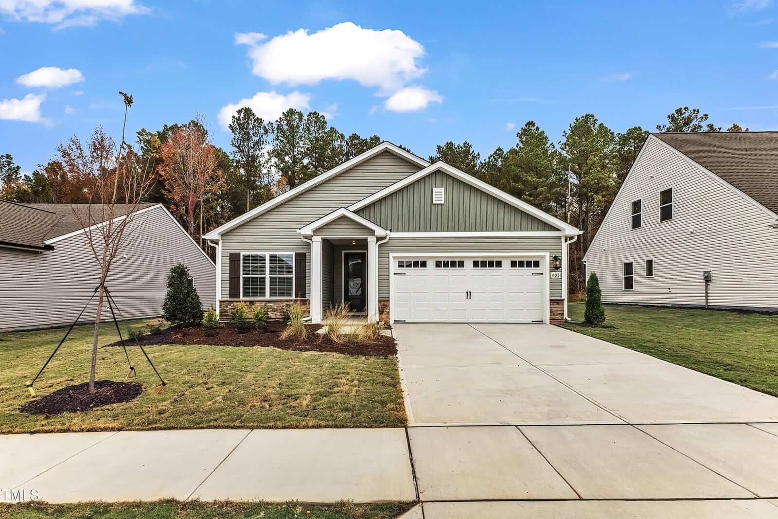 Single-story house with green and white exterior, two-car garage, covered front porch, and neatly landscaped yard with grass, small trees, and shrubs. Concrete driveway and sidewalk in a suburban neighborhood.