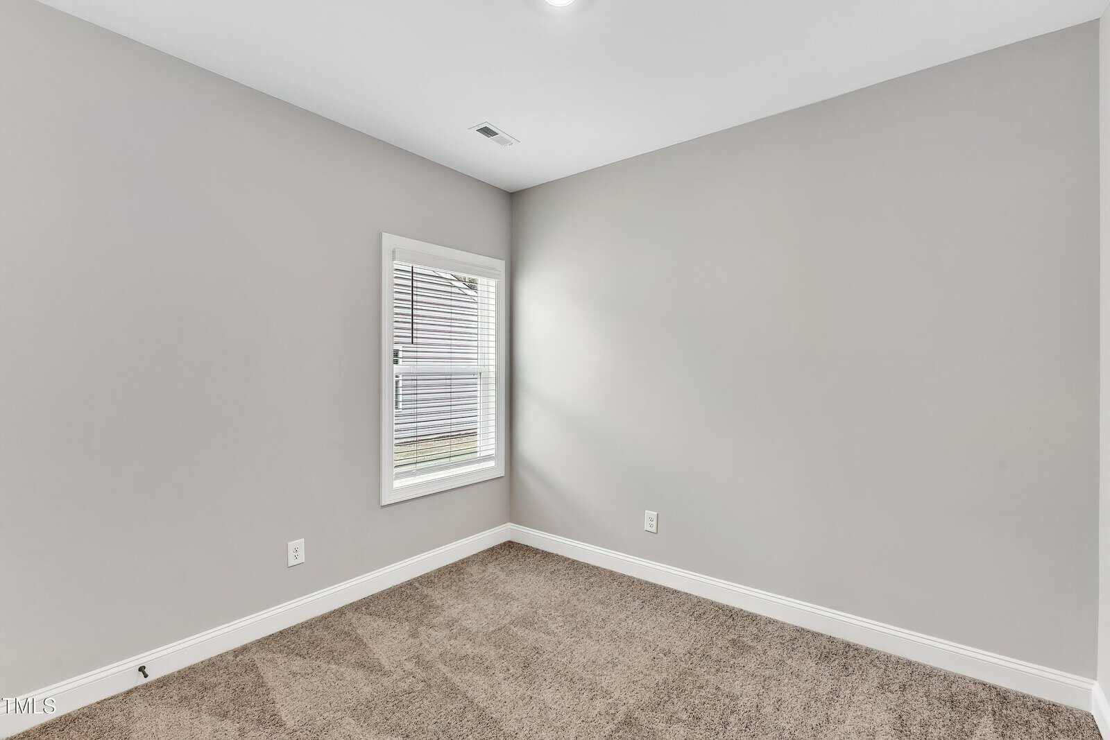 A small, empty room with light gray walls, a window with white blinds, white trim, and beige carpet flooring. Natural light comes through the window, creating a bright and clean appearance.